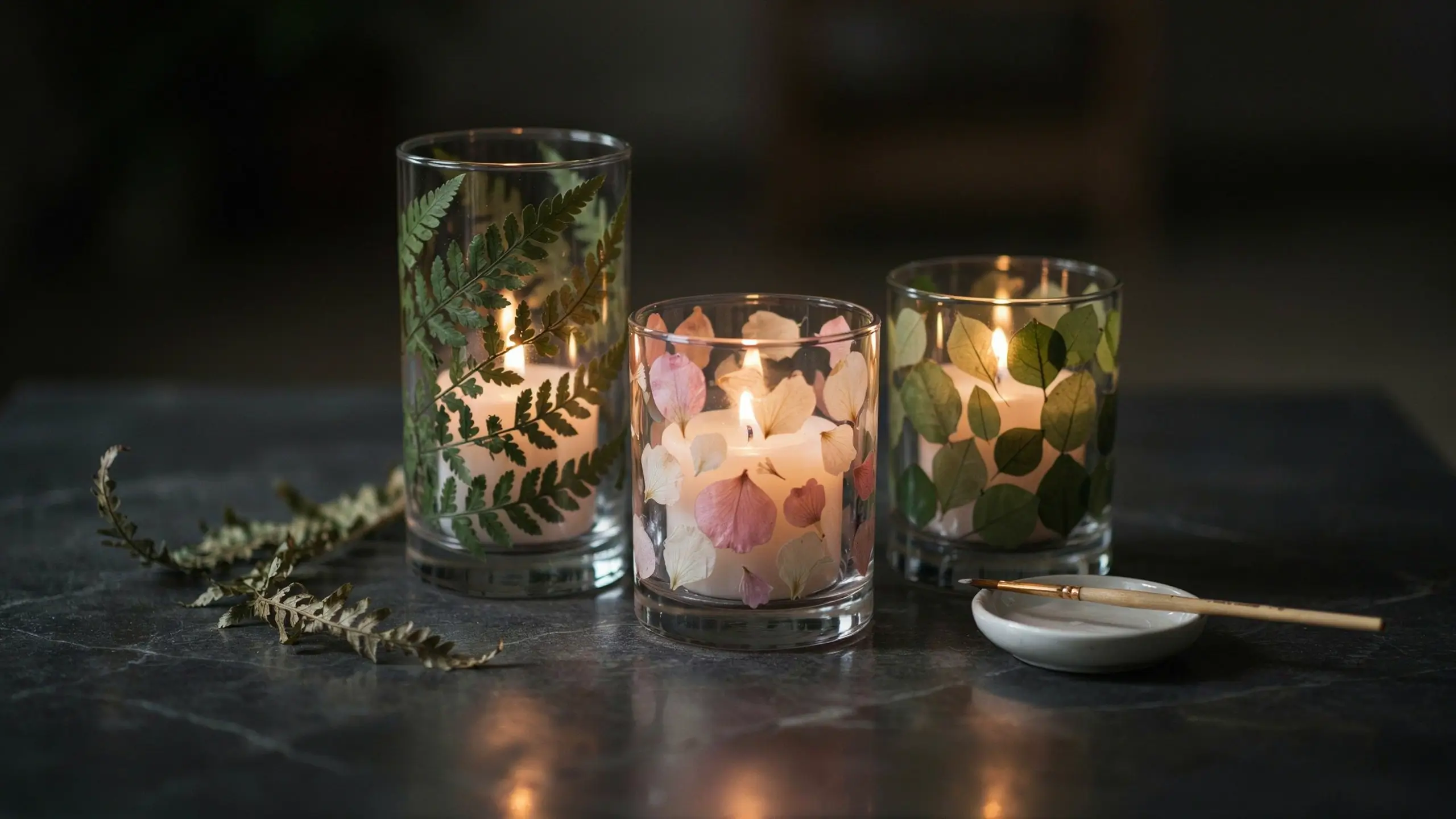 Three glass jar candle holders decorated with pressed ferns and pink hydrangea petals, glowing with lit white candles on a dark slate table for a spring DIY project. 
