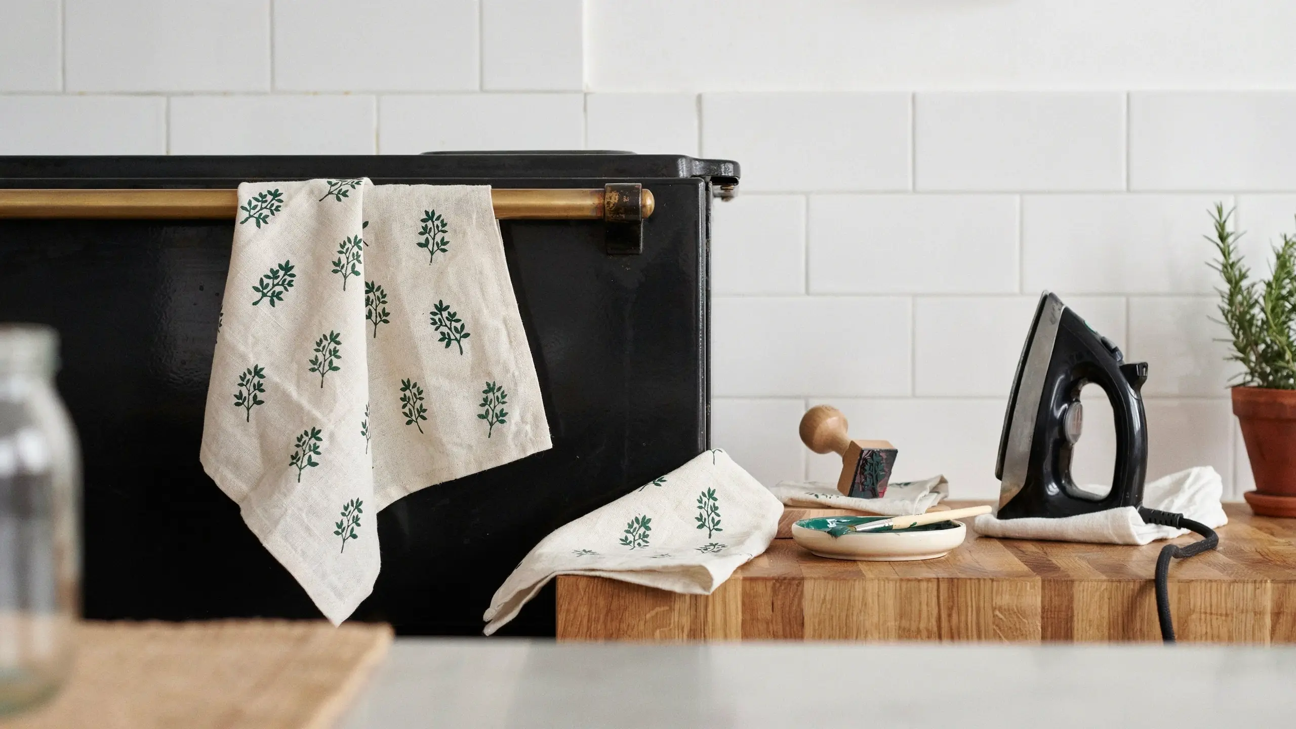 Hand-stamped white linen tea towels with green leaf motifs, shown with a wooden stamp, ink dish, and iron on a wooden counter. 
