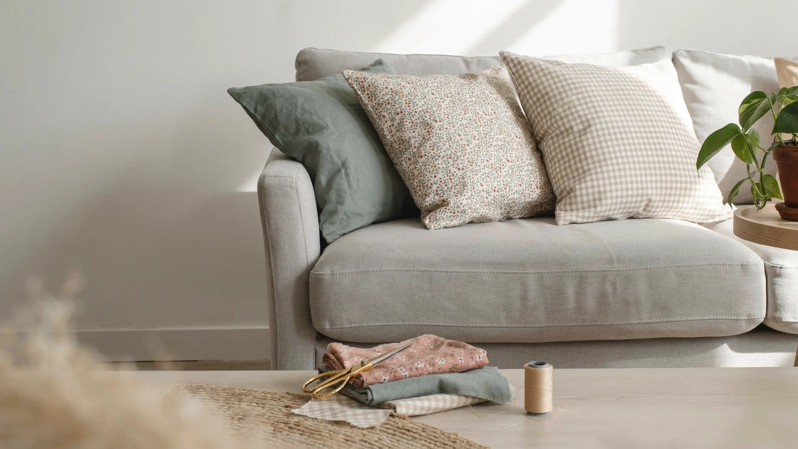 Light grey sofa with floral and gingham DIY pillow covers, with thrifted fabric, scissors, and thread on a table for budget sewing swap. 