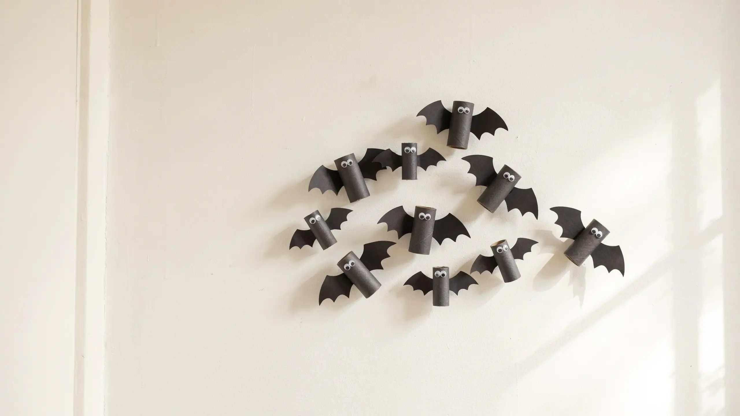 Close-up of a group of handmade Halloween bat decorations crafted from black painted toilet paper rolls with construction paper wings and googly eyes, mounted on a white wall as a budget-friendly kids craft.