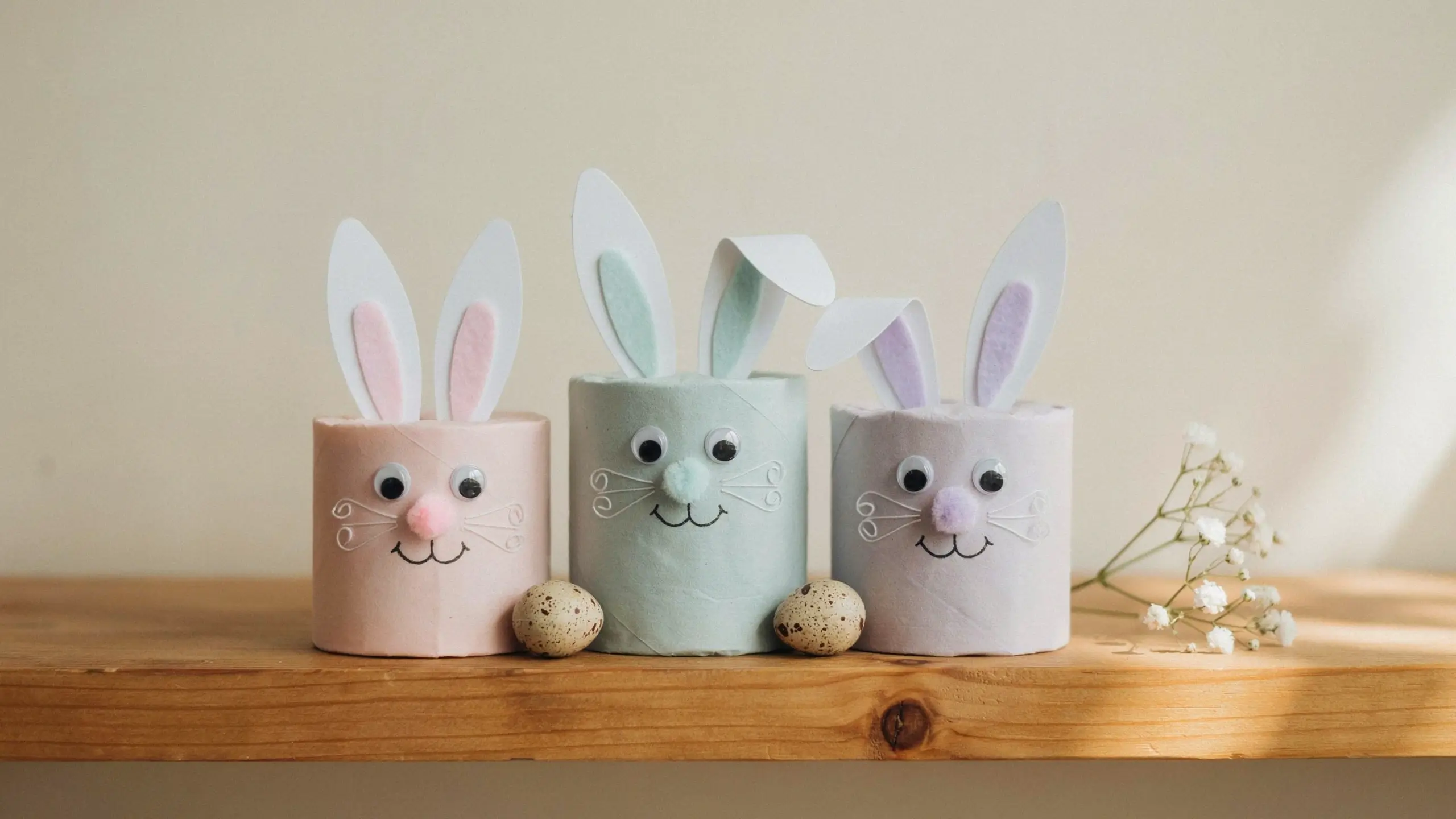 Three pastel painted DIY toilet paper roll bunnies with googly eyes and felt ears, displayed with speckled Easter eggs on a wooden shelf. 