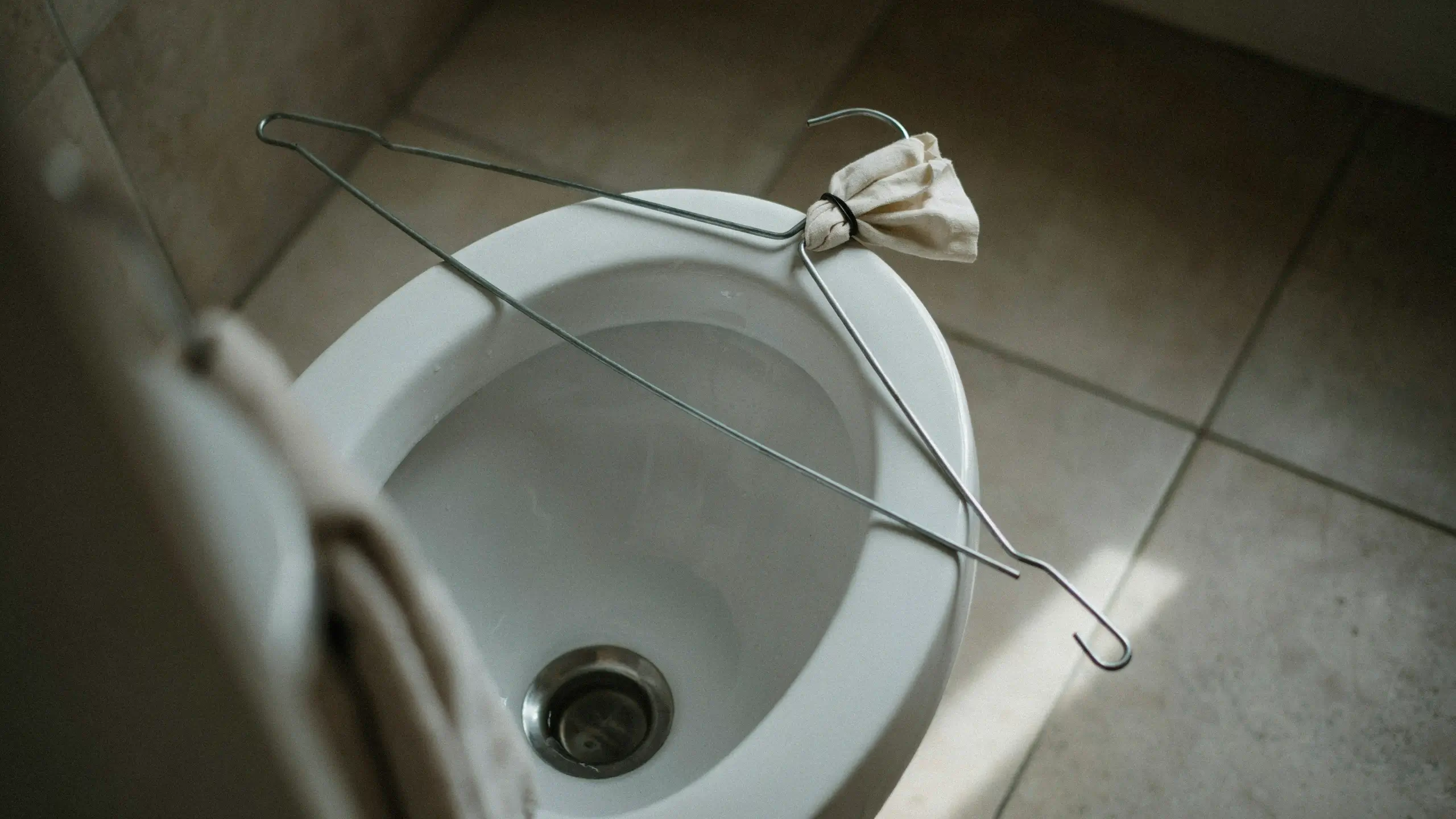 Wire coat hanger with a tied rag resting across the rim of a white ceramic bathroom sink, used as a makeshift DIY plumbing tool for clearing drains