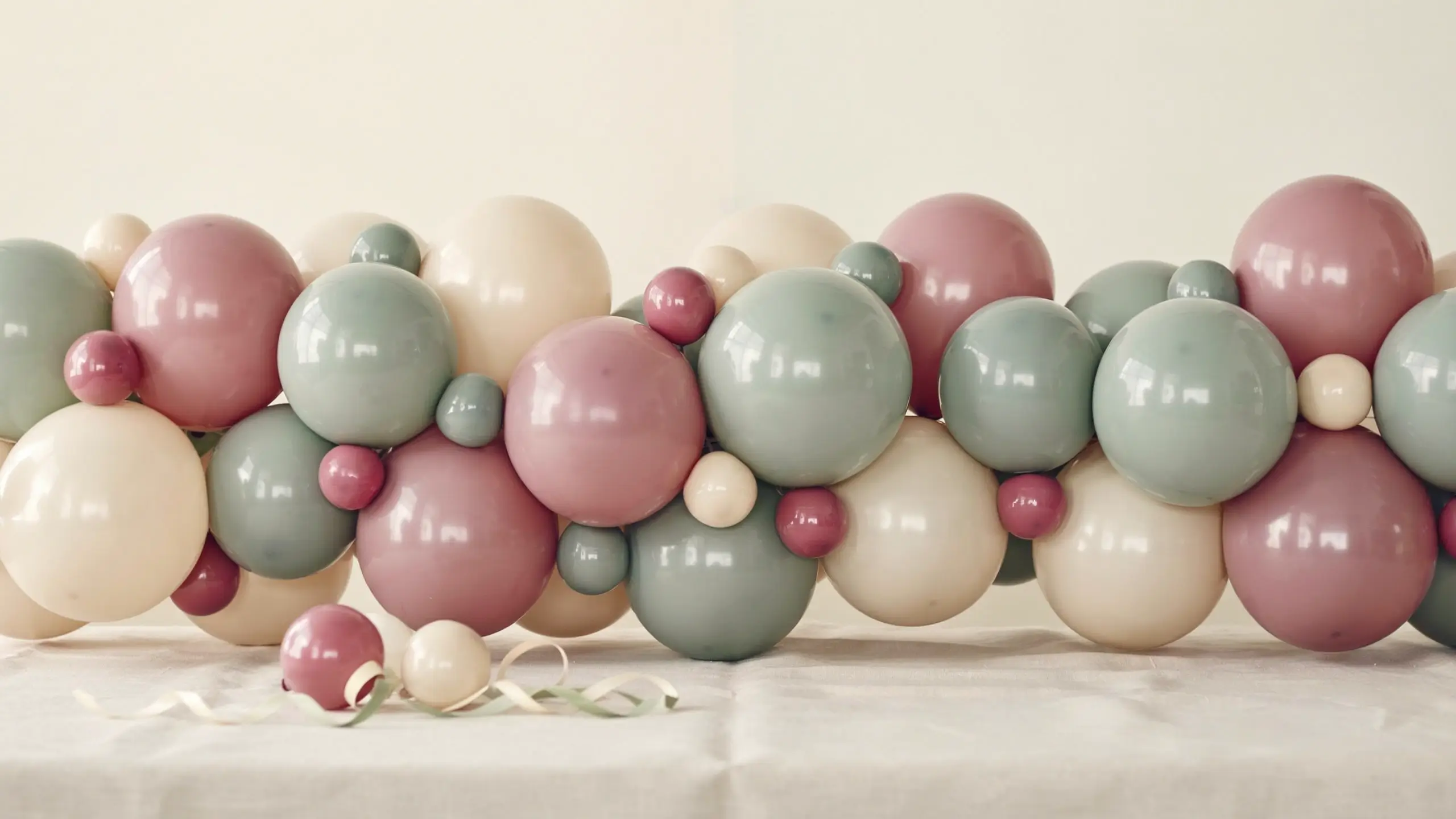 Dusty rose, sage green, and cream balloon garland arranged on a white tablecloth, featuring small berry-colored mini balloons used to fill gaps and add texture between the larger balloons for a cohesive party setup.