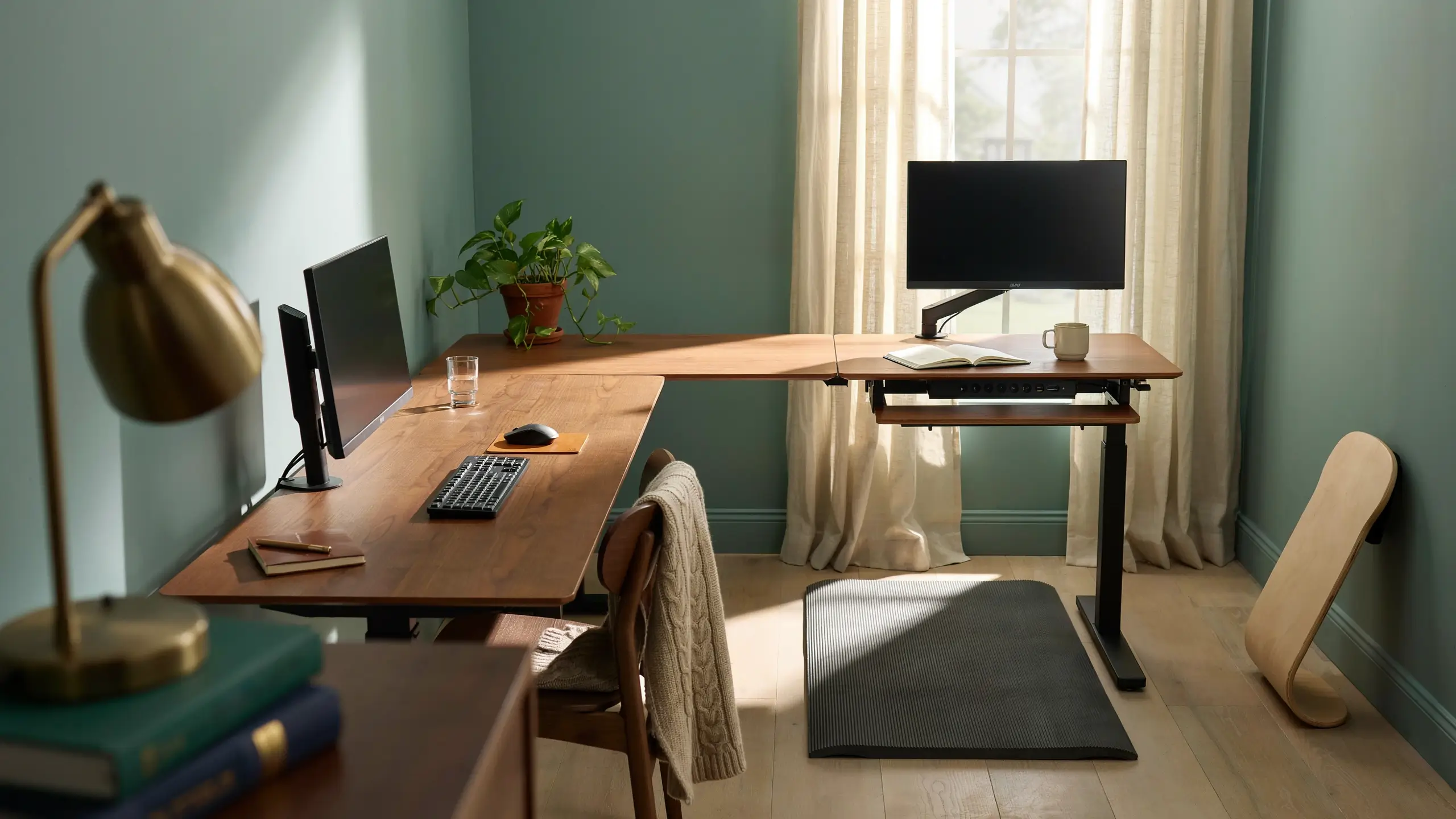 L-shaped standing desk in a sunlit home office featuring dual monitors, a keyboard tray, an anti-fatigue floor mat, and a potted plant against a sage green wall. 