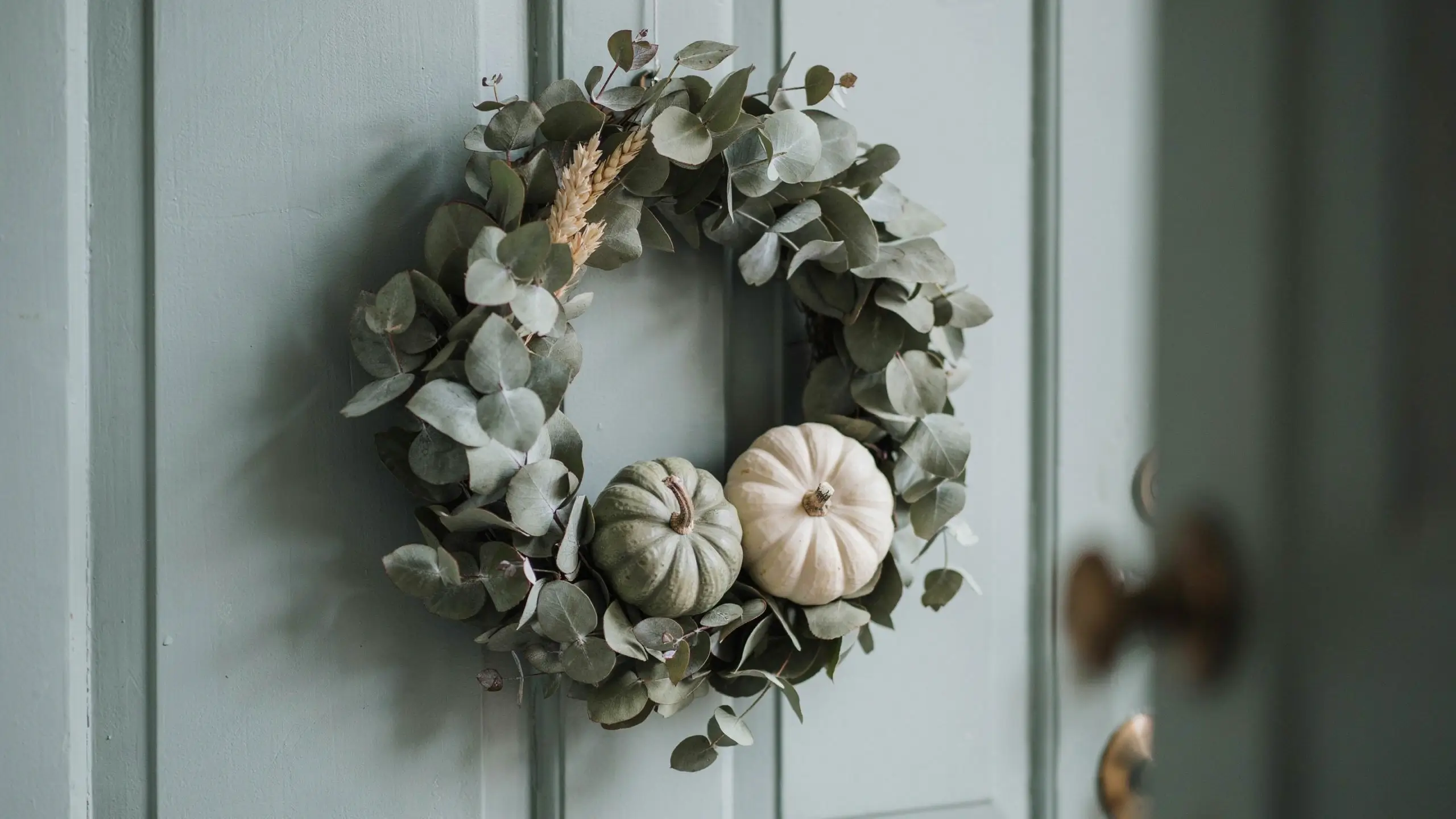 Sage green eucalyptus wreath with a white and green mini pumpkin and wheat stalks hanging on a light teal front door.