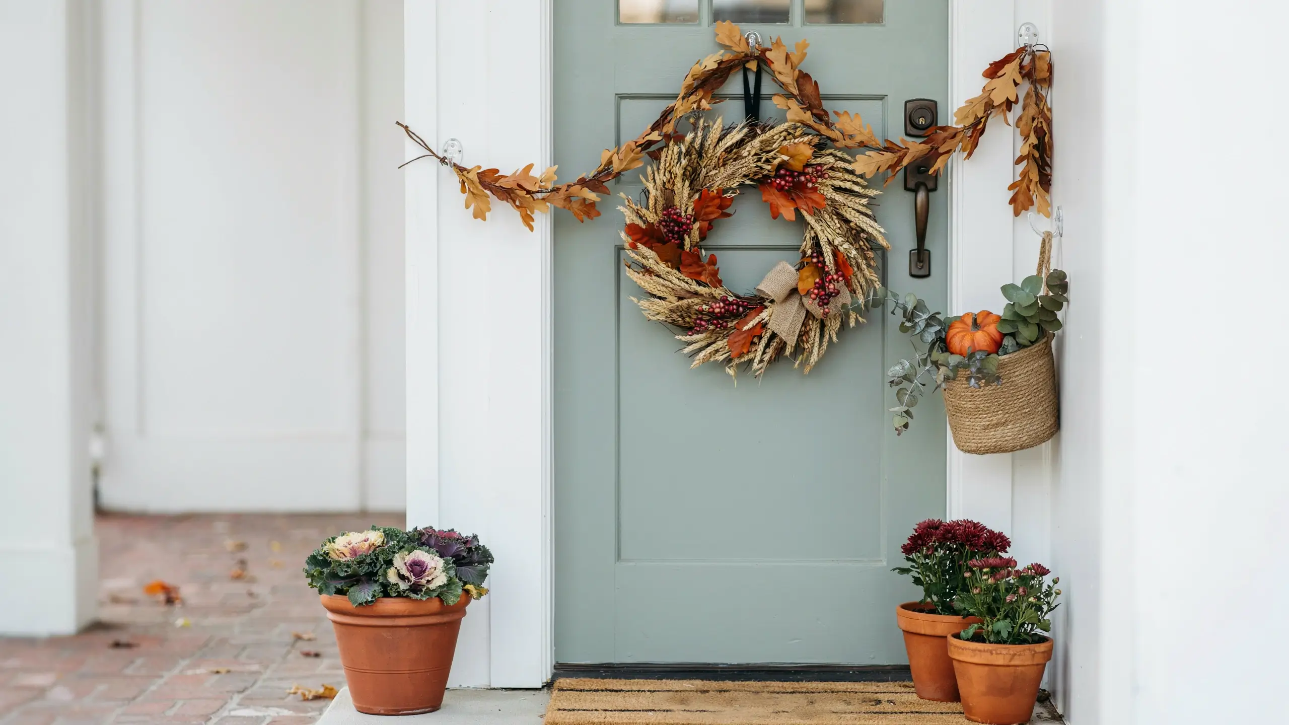 Alt Text: Sage green front door decorated with an autumn wheat wreath, leaf garland, potted mums, and a small pumpkin.