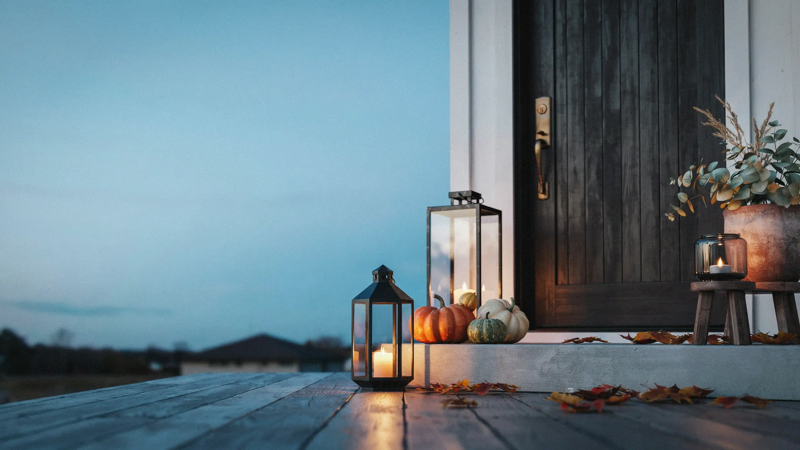 Alt Text: Fall front porch with glowing lanterns, pumpkins, and autumn leaves beside a black door at dusk.