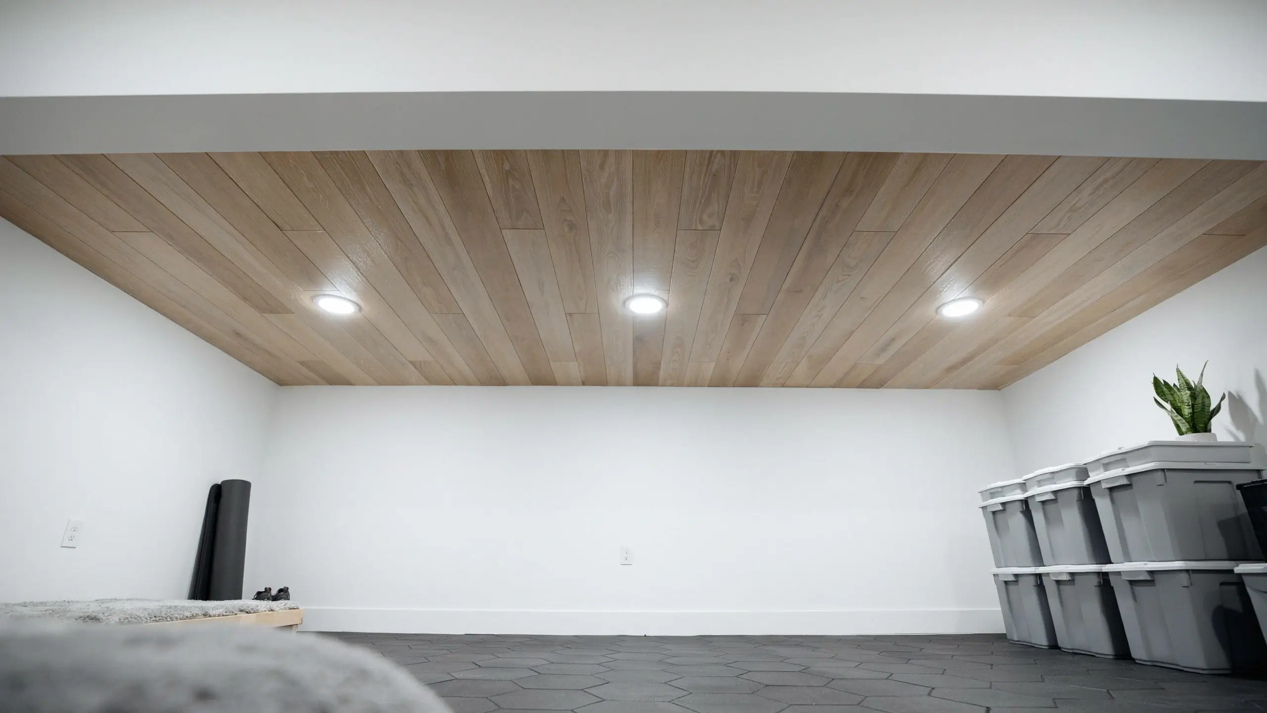 A sloped room featuring a light faux wood panel ceiling with three recessed lights, white walls, dark grey hexagonal floor tiles, and grey storage bins to the right.
