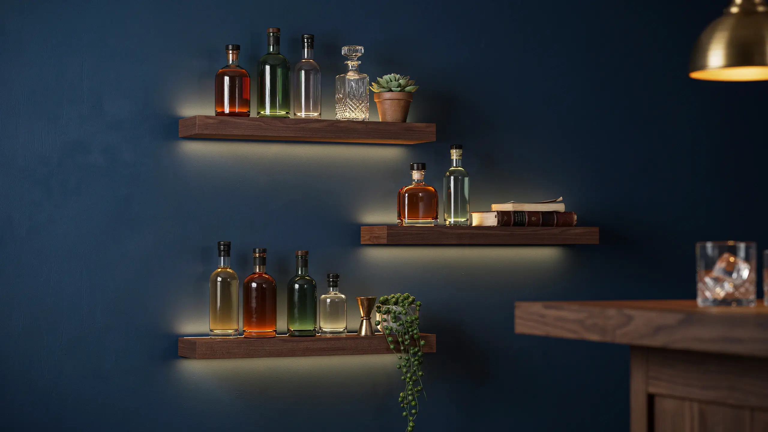 "Three dark walnut floating wooden shelves mounted on a deep navy blue accent wall, displaying a curated collection of amber and clear liquor bottles, a crystal decanter, small potted succulents, a brass jigger, and stacked books, illuminated by warm ambient lighting in a modern home bar setup." 