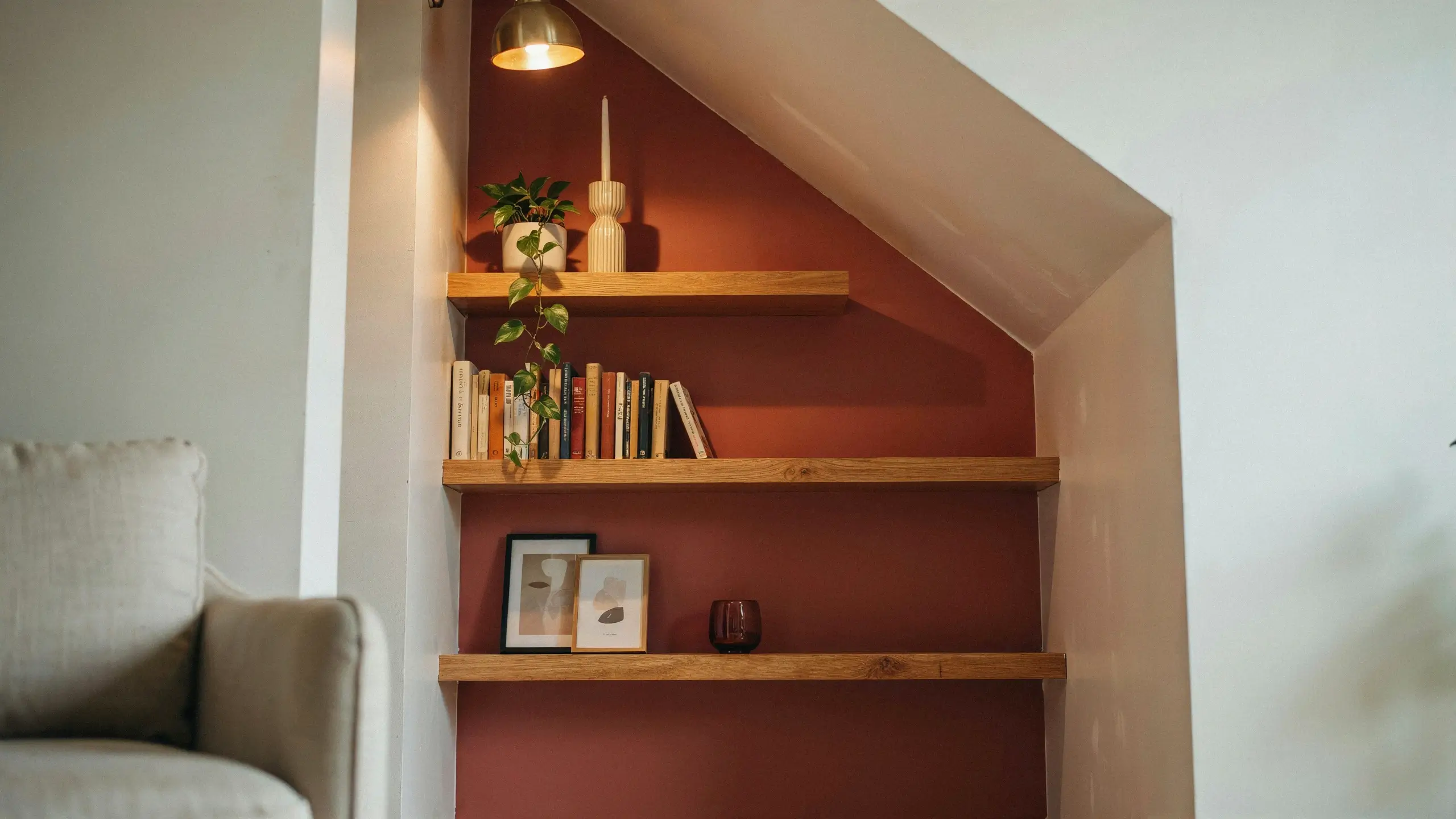 Three tiered oak floating shelves displaying stacked books, framed abstract art, and a trailing pothos plant, mounted inside a home alcove painted with a matte terracotta accent wall and illuminated by a warm brass pendant lamp.