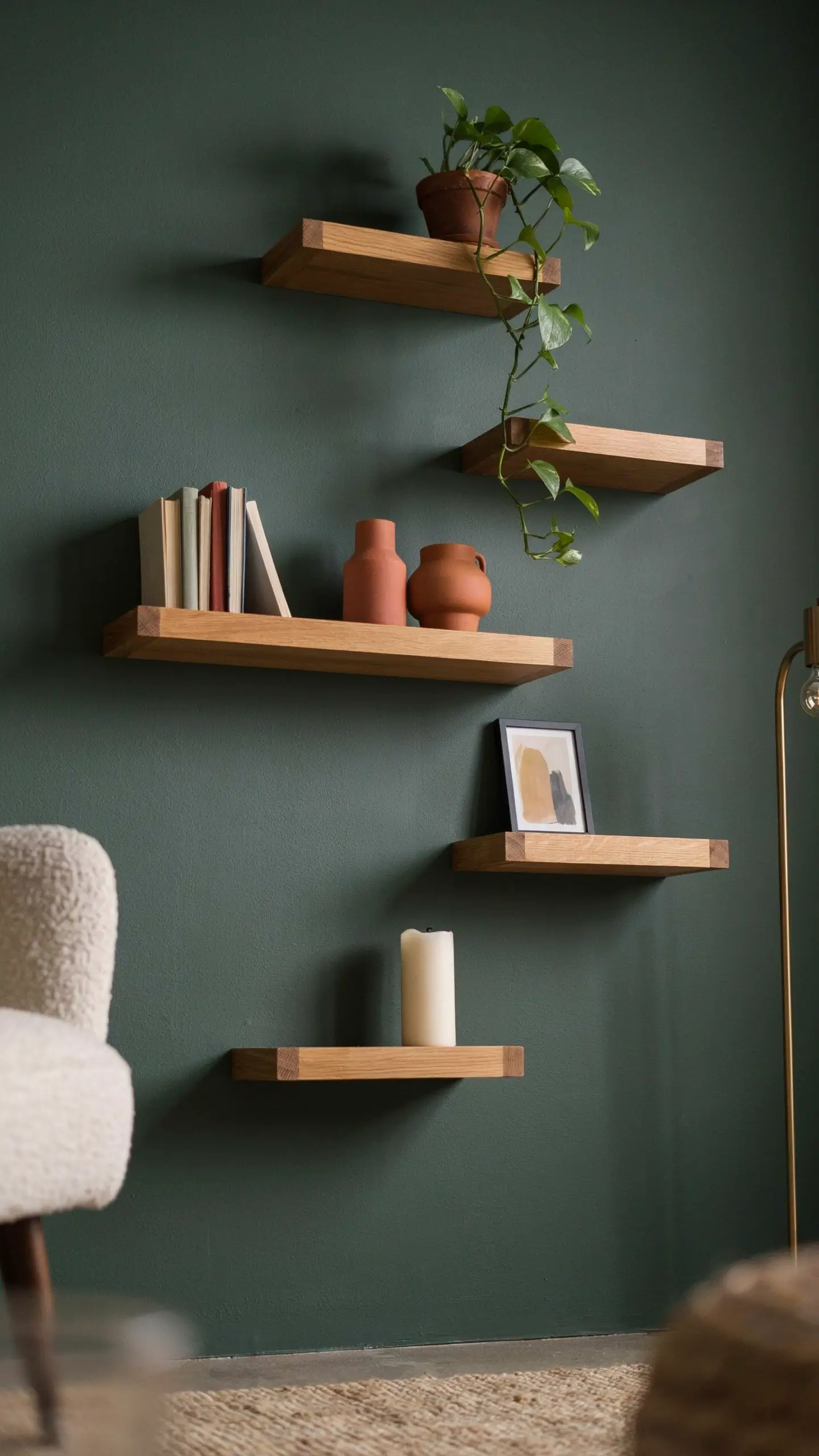 "Image of five natural wood floating shelves arranged on a dark matte green accent wall, decorated with a trailing pothos plant, terracotta pottery, modern books, and minimalist framed art in a cozy living room corner." 