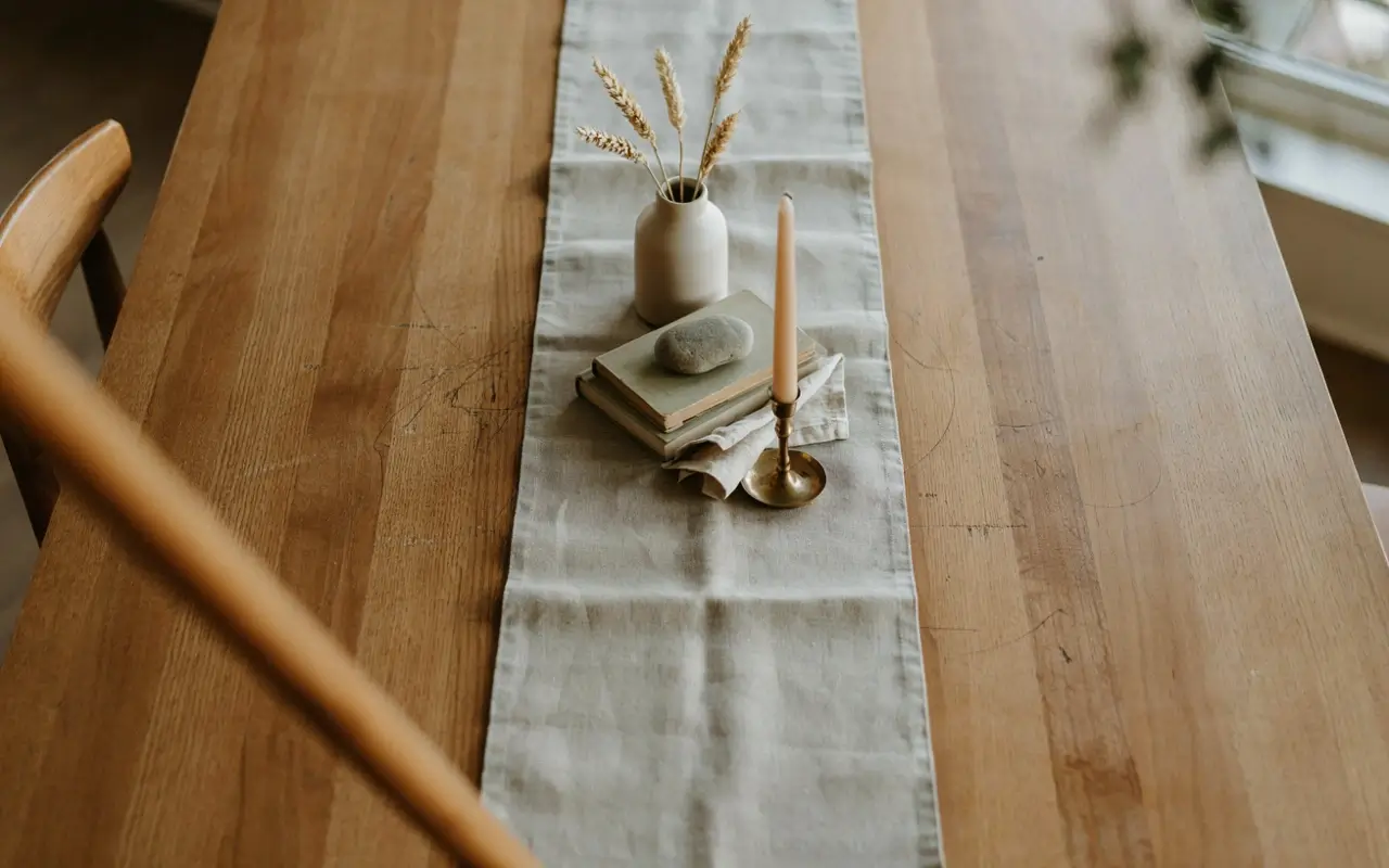 Folded linen runner on a wood dining table with a small vase, stacked books, stone, and brass taper candle.