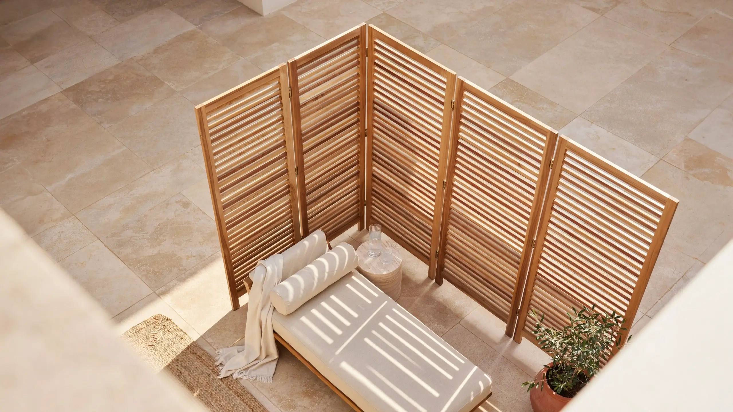 Top-down view of a freestanding wood slatted accordion folding screen arranged in a corner formation to create a private, sunlit lounging space with a daybed, side table, and potted plant. 