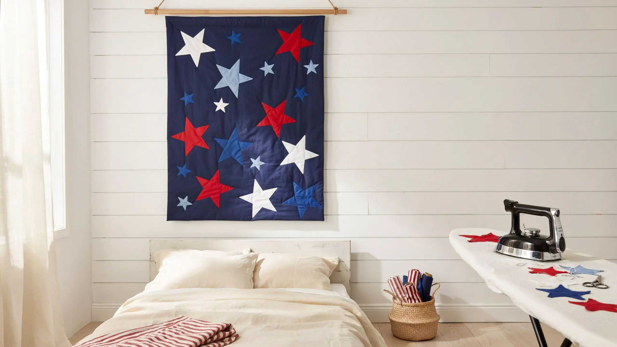 Navy blue fused star cluster appliqué wall quilt hanging in a rustic white shiplap bedroom with an ironing board holding a chrome steam iron and precut fabric stars