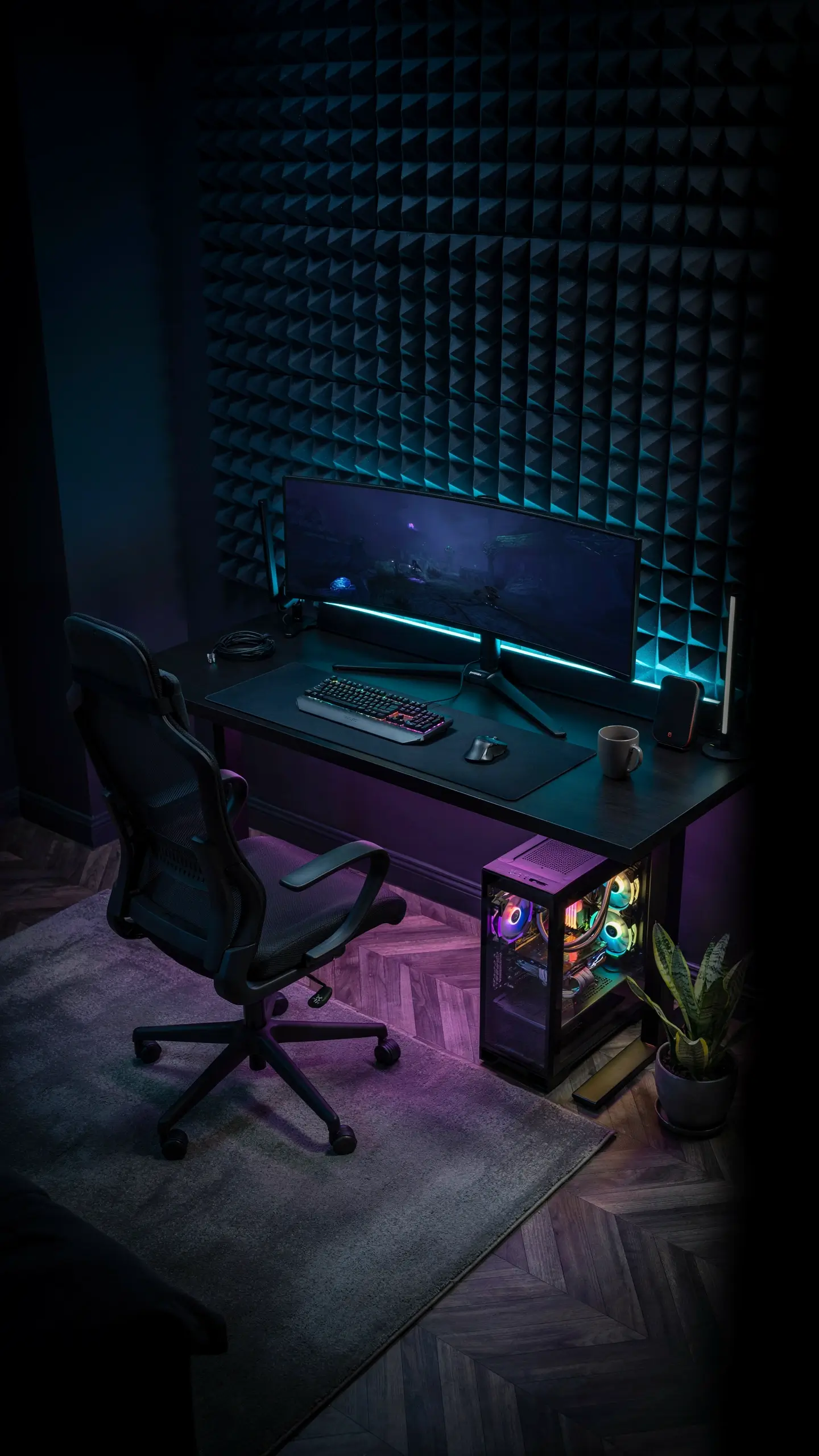 A moody dark gaming setup featuring an ultrawide monitor, glowing RGB keyboard, and glass PC tower, placed against a black textured acoustic foam wall with cyan and purple ambient LED backlighting. 