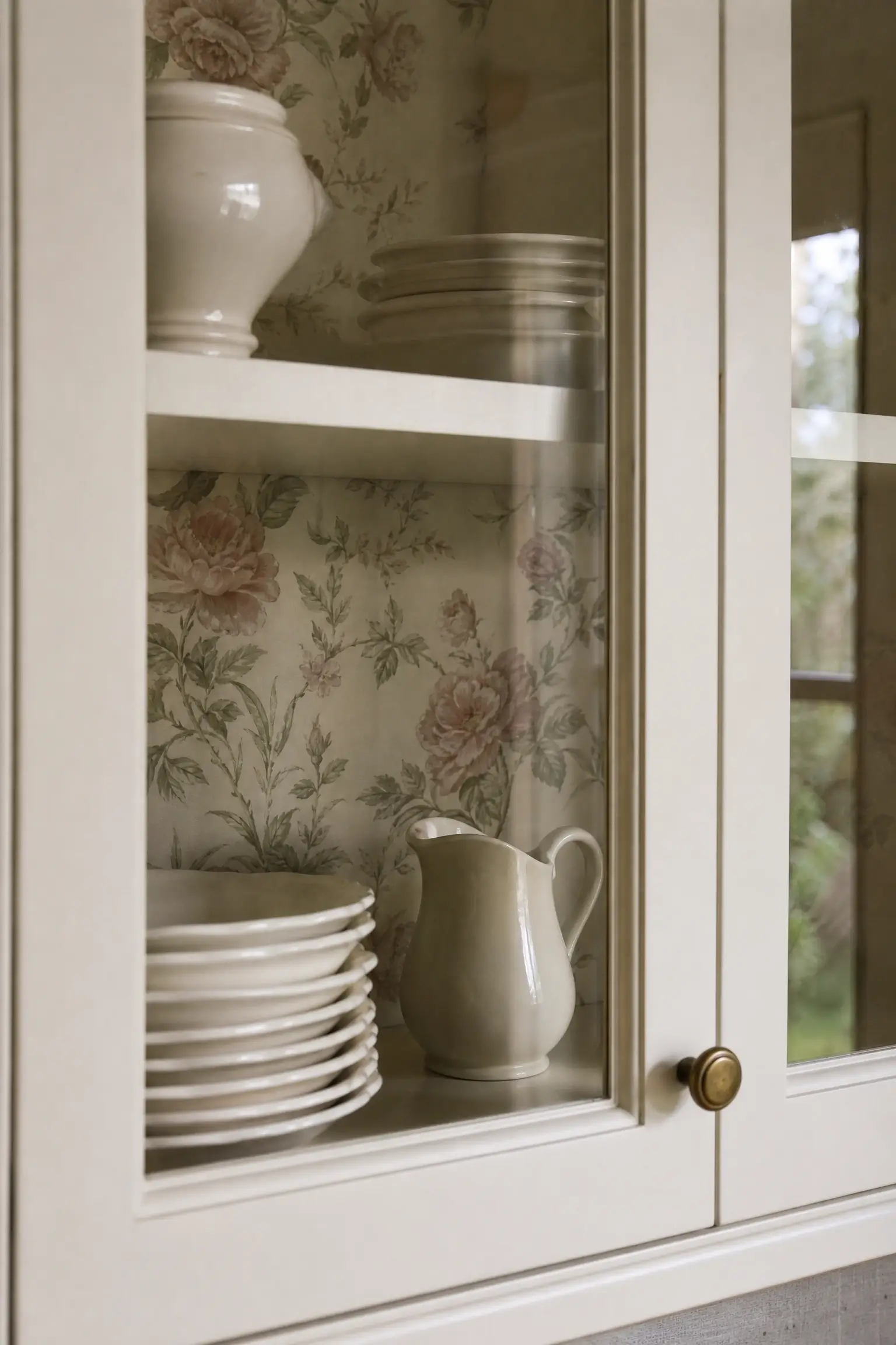 Glass-front kitchen cabinet with floral wallpaper backing, cream plates, and a ceramic pitcher.