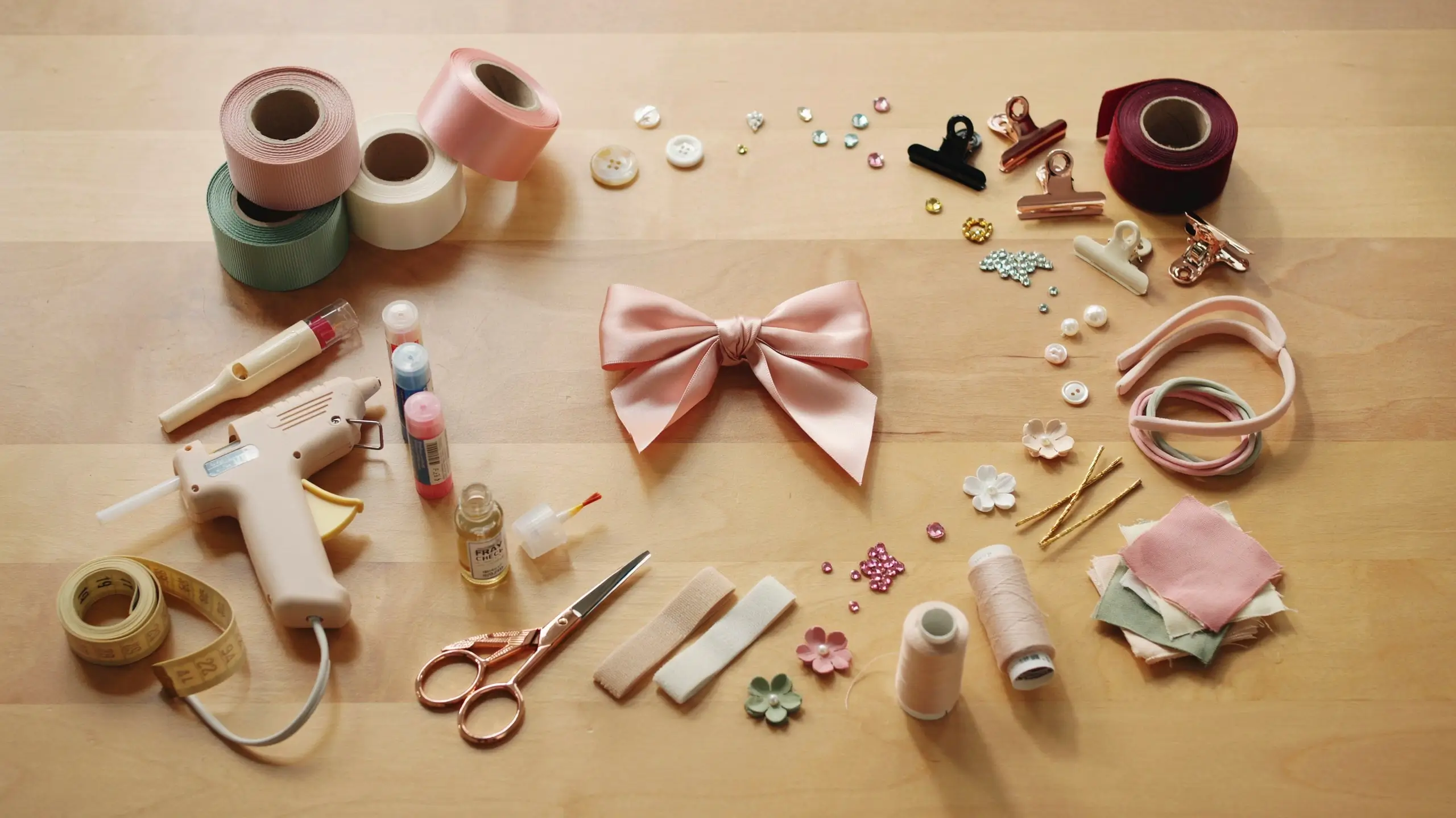 Alt Text: Hair bow making supplies with ribbon, scissors, glue gun, clips, thread, and a finished pink bow