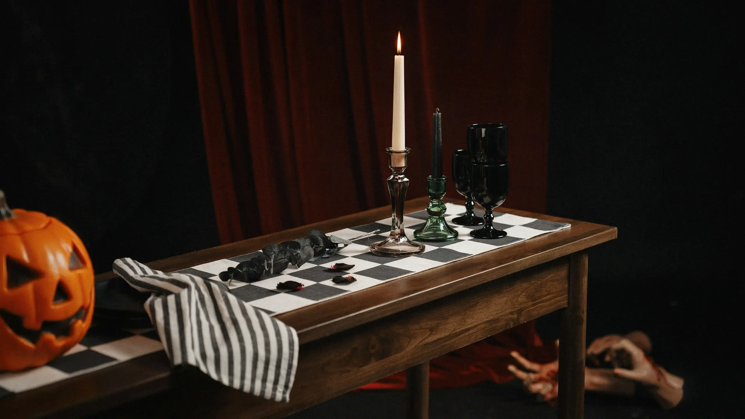 A spooky Halloween table setting featuring an orange carved jack-o-lantern, a black and white checkered runner, a tall taper candle in a silver holder, black goblets, and creepy hands reaching out from under the wooden table against a dark red curtain backdrop.