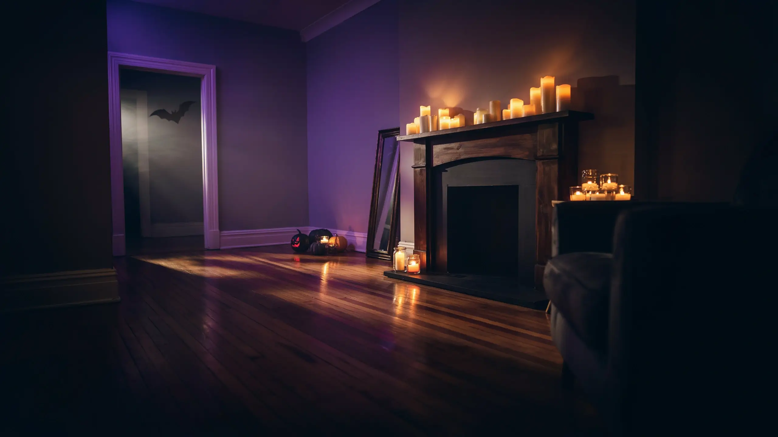 "Dimly lit Halloween living room decor featuring a wooden fireplace topped with glowing pillar candles, purple ambient wall lighting, a bat silhouette on the open doorway, and small jack-o'-lanterns on the reflective wooden floor."