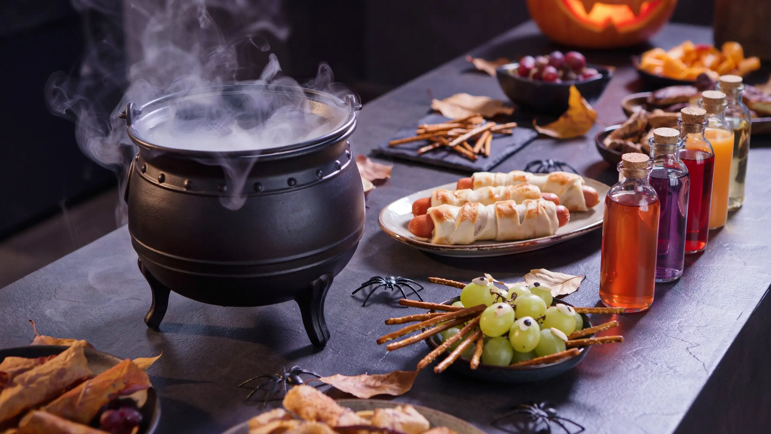 Halloween party food spread featuring a steaming black cauldron, mummy-wrapped hot dogs, and green grape eyeballs with pretzel sticks.