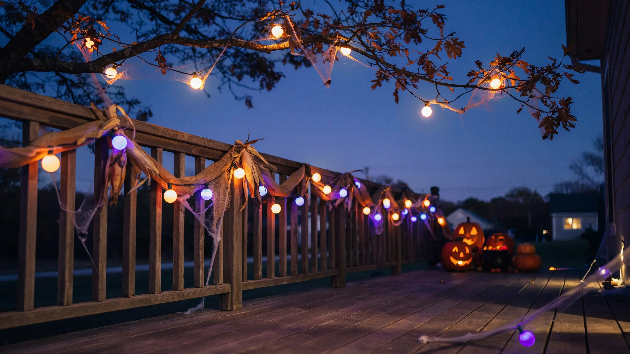 "Wooden deck decorated for Halloween with alternating glowing orange and purple string lights, dried corn husks, fake spiderwebs, and illuminated jack-o'-lanterns set against a twilight blue sky." 