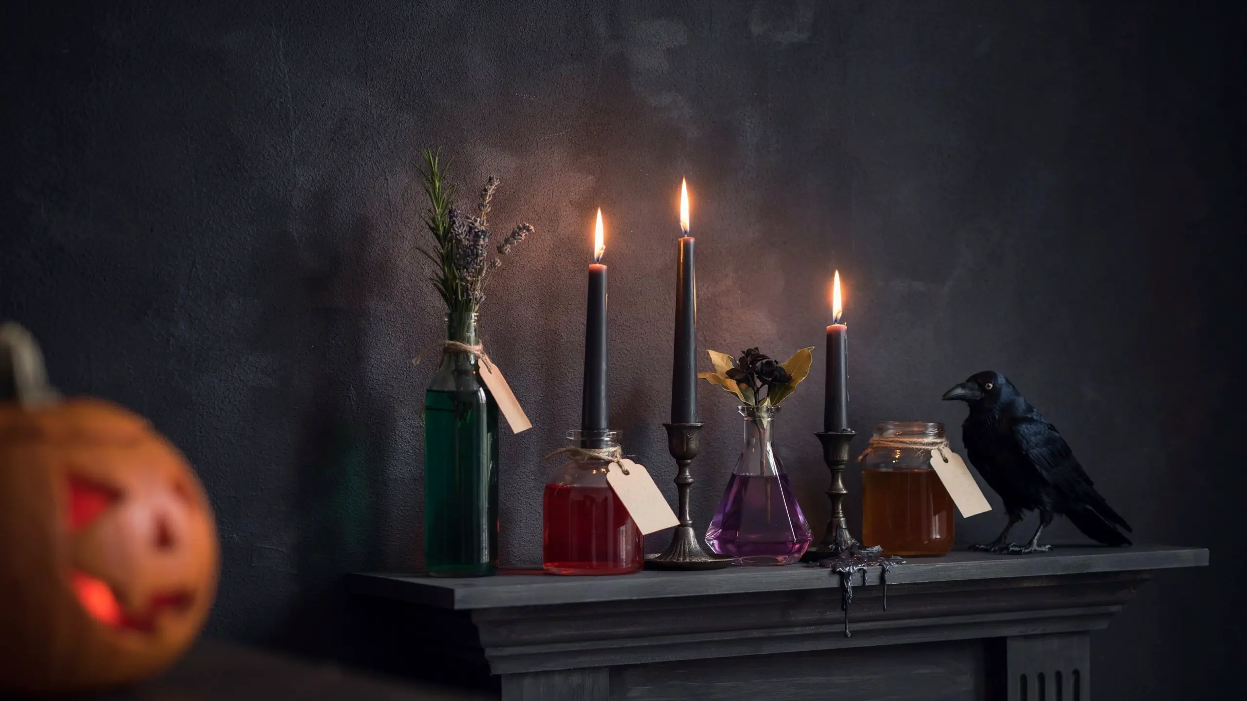 A moody Halloween mantel display featuring glass apothecary jars filled with glowing red, purple, green, and amber potion liquids. Three lit black taper candles in ornate metal holders, a realistic black crow prop, a carved orange jack-o'-lantern, and tied rustic paper labels sit against a dark, textured charcoal wall. 