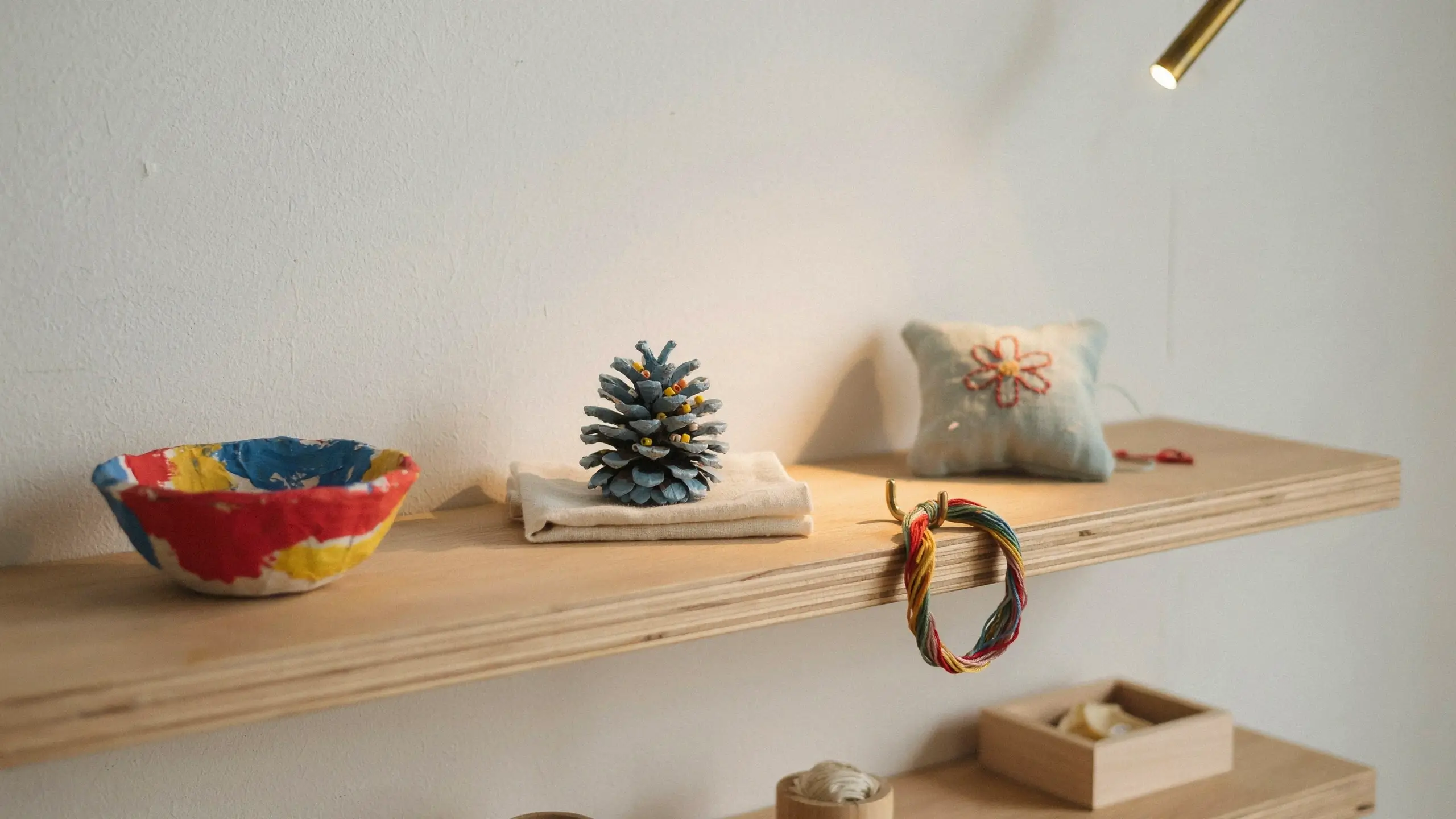 A curated wooden shelf displaying finished DIY craft projects including a patterned painted bowl, a blue painted pinecone on a white cloth, a hand-embroidered floral pillow, and a colorful yarn ring hanging from the edge, illuminated by warm accent lighting. 