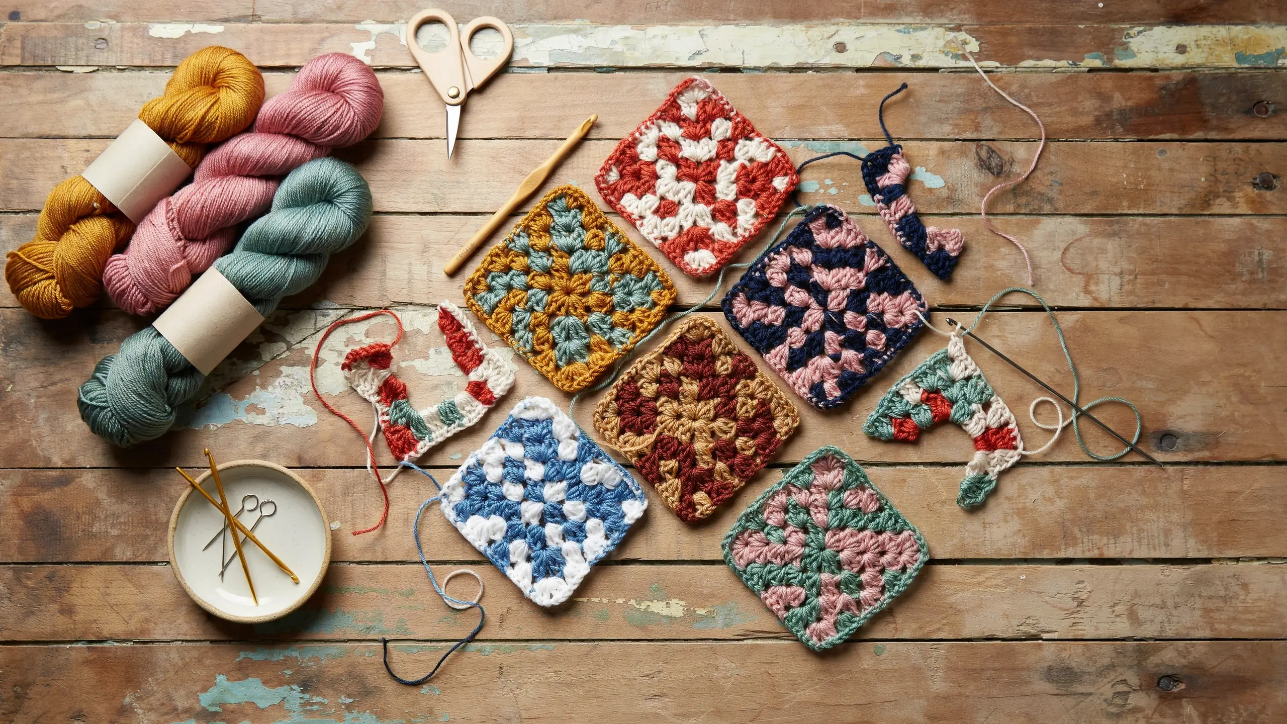 A flat lay of colorful handmade crochet granny squares paired with skeins of pastel and mustard yarn, a wooden crochet hook, small scissors, and sewing needles resting on a rustic wooden background.