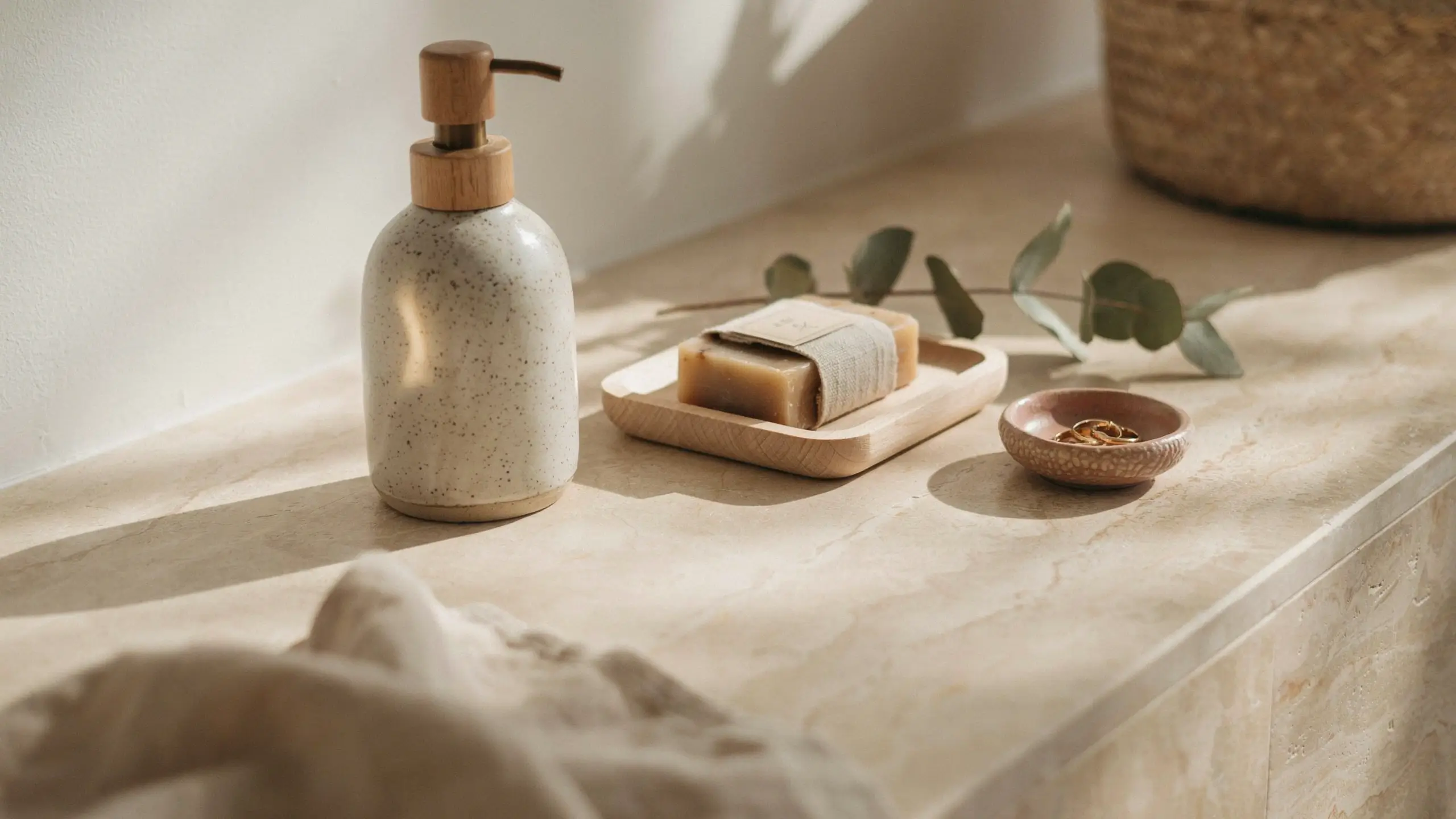 Handmade speckled ceramic soap dispenser with a bamboo pump next to a natural wooden soap tray holding a bar of soap, resting on a light beige marble bathroom counter. The spa-like scene includes a eucalyptus branch, a small terracotta dish with gold rings, and soft natural sunlight casting strong shadows.