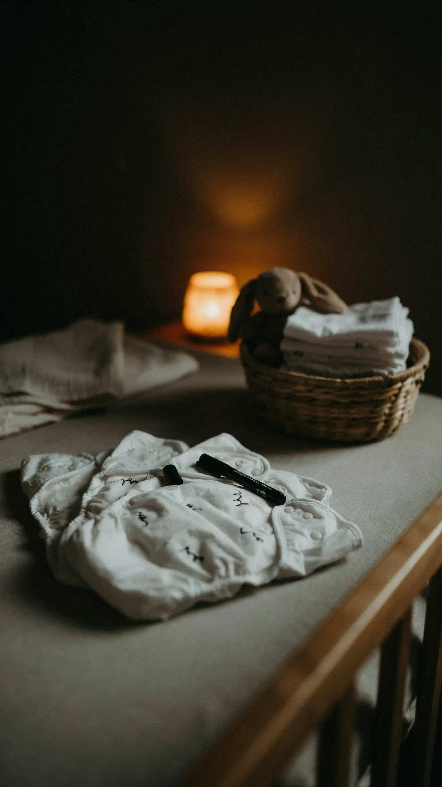 White reusable cloth diaper with handwritten messages and markers on a changing table, next to a basket of folded diapers, a plush bunny, and a warm candle in a dim nursery. 
