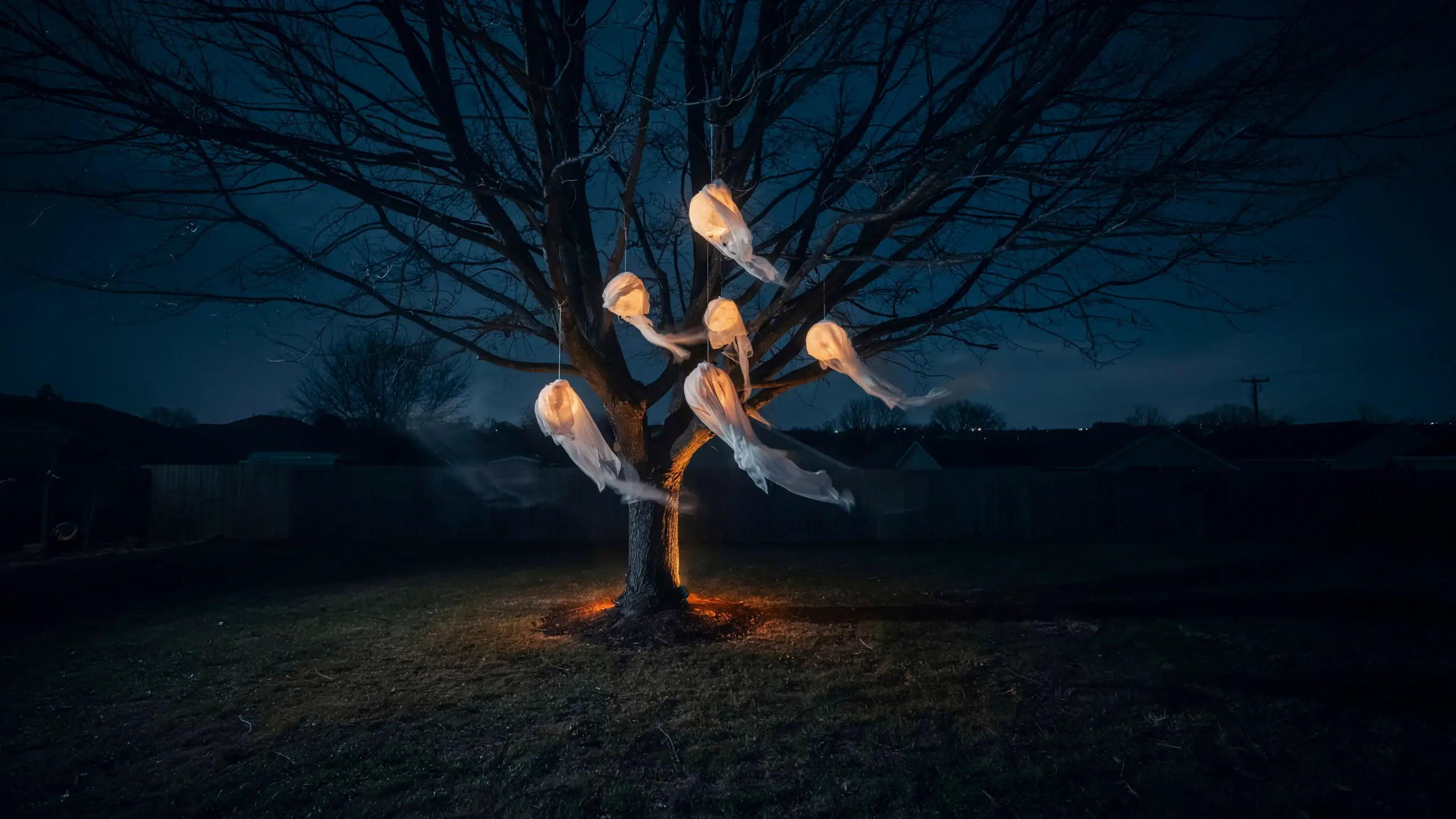 A cluster of glowing, white DIY cheesecloth ghosts hanging from the bare branches of a large tree at night, illuminated by warm orange uplighting to create a spooky backyard Halloween display.