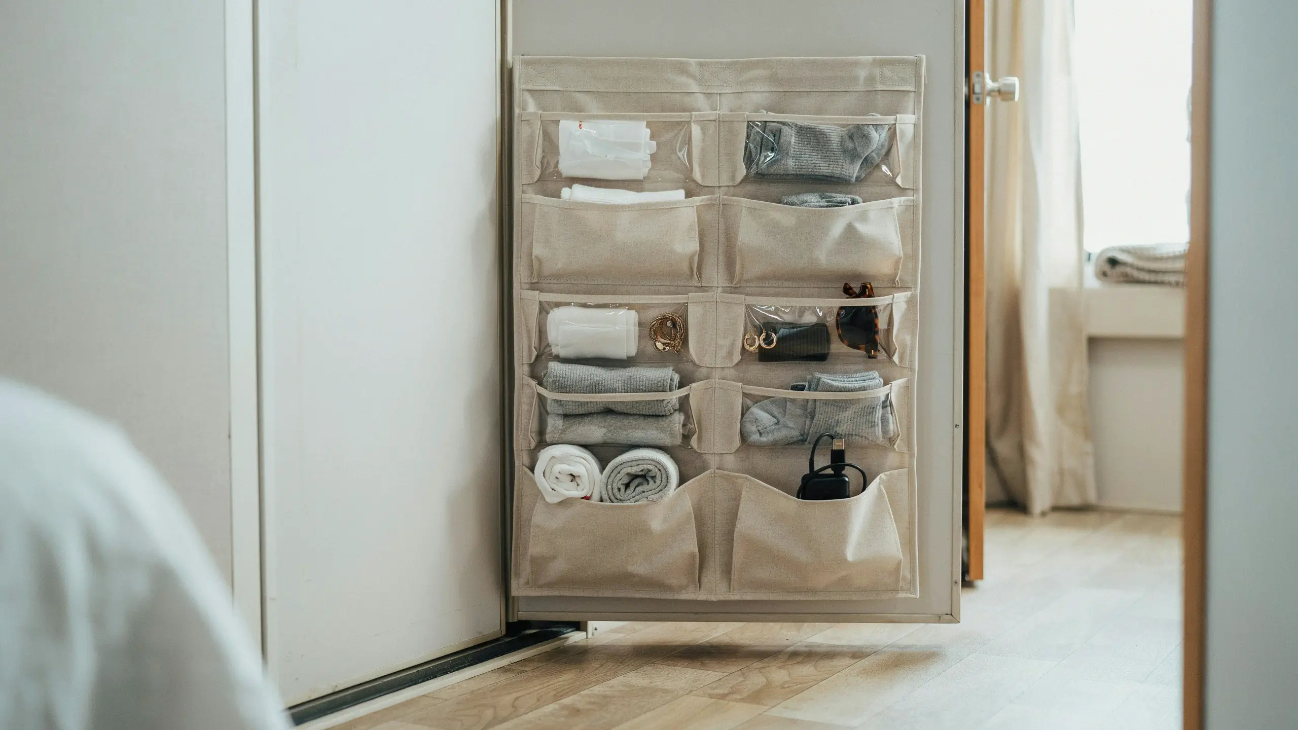 Beige fabric over-the-door hanging organizer with multi-sized clear pockets storing rolled towels, sunglasses, jewelry, and electronic chargers inside a white closet
