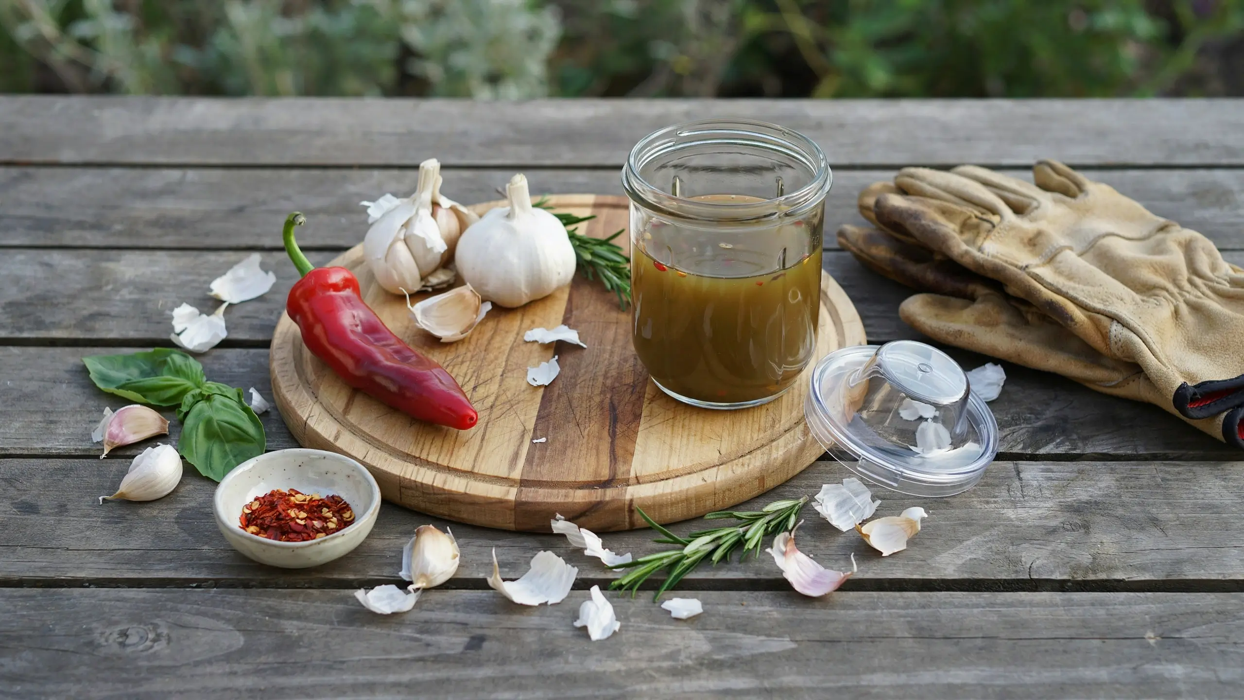 DIY garlic and pepper spray ingredients on a wooden cutting board, including garlic bulbs, red chili, chili flakes, fresh rosemary, and a glass jar of infused oil for natural garden pest control. 