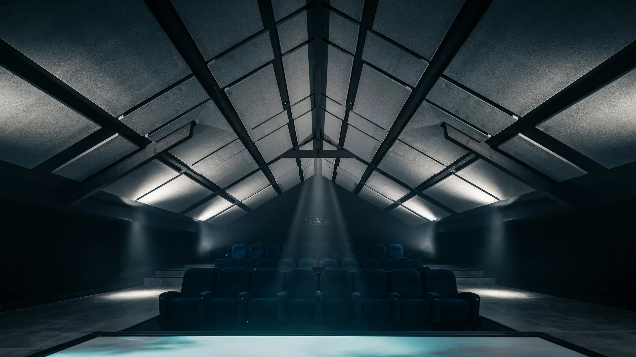 Modern home theater featuring tiered dark stadium seating, dramatic spotlight beams, and a vaulted ceiling with acoustic panels and exposed structural beams