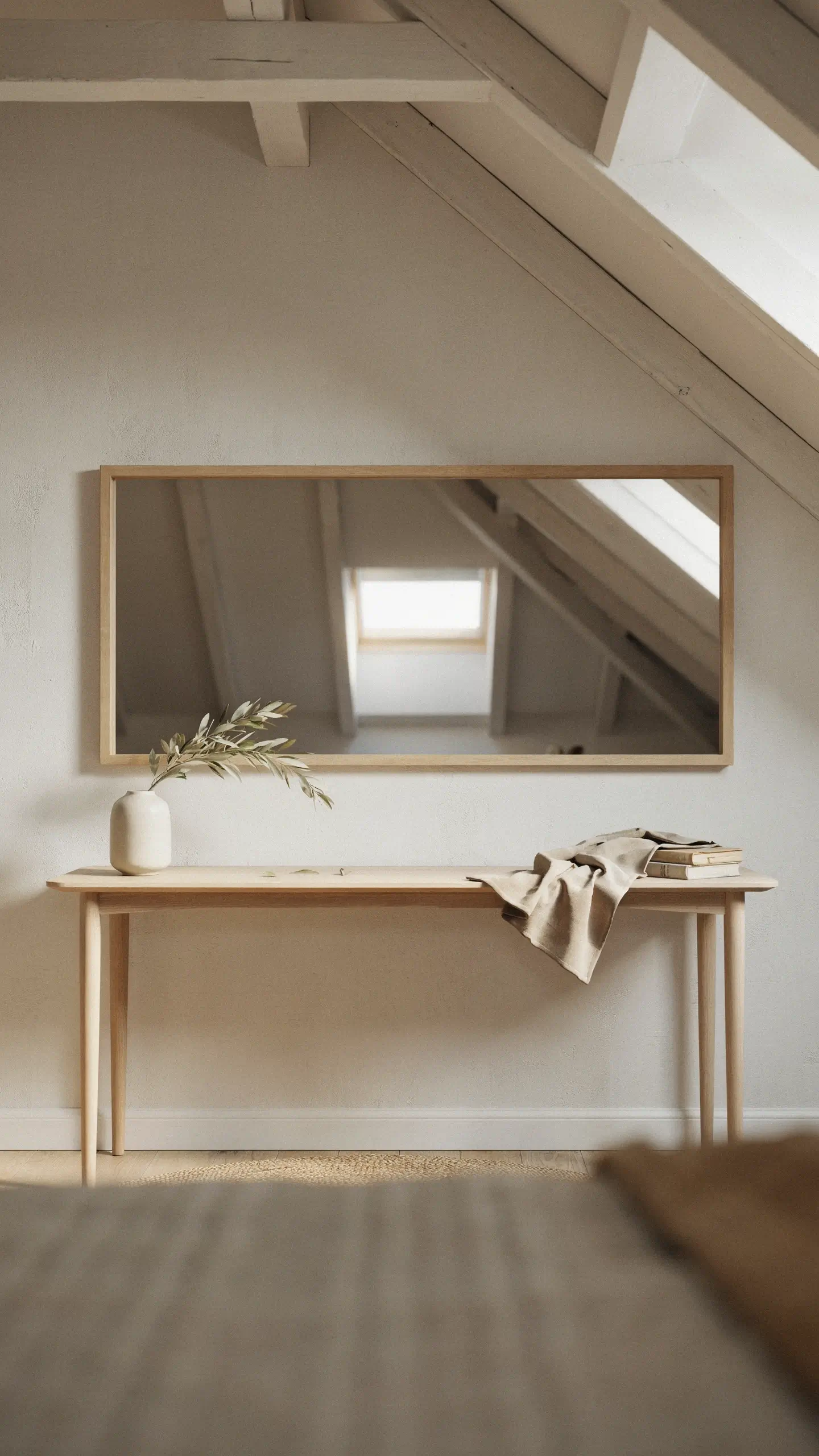 Horizontal rectangular mirror with a light wood frame mounted on a white attic wall, reflecting a skylight window and slanted ceiling beams, situated above a minimalist light wood console table with a ceramic vase, books, and a draped beige cloth. 