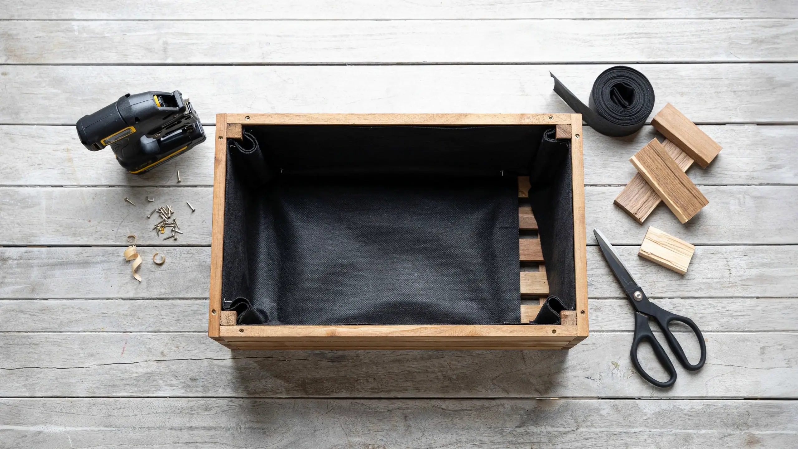 Overhead view of a DIY wooden planter box being lined with black landscape fabric, alongside a cordless drill, scissors, screws, and scrap wood blocks resting on a white wooden workbench.