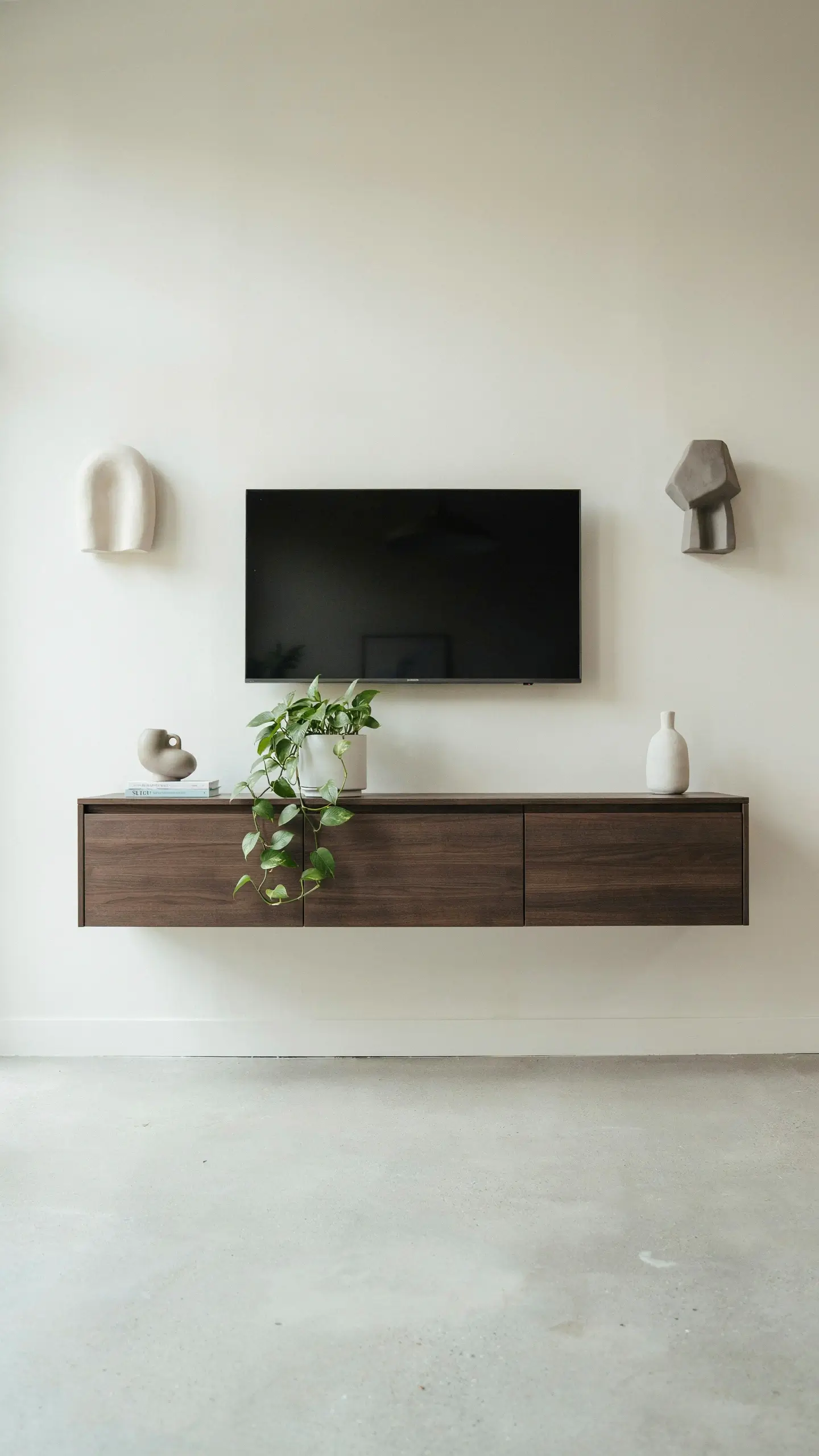 Dark walnut wood BESTÅ floating media console mounted on an off-white wall, styled with a cascading pothos plant, abstract ceramic vases, and geometric wall art in a minimalist living room. 