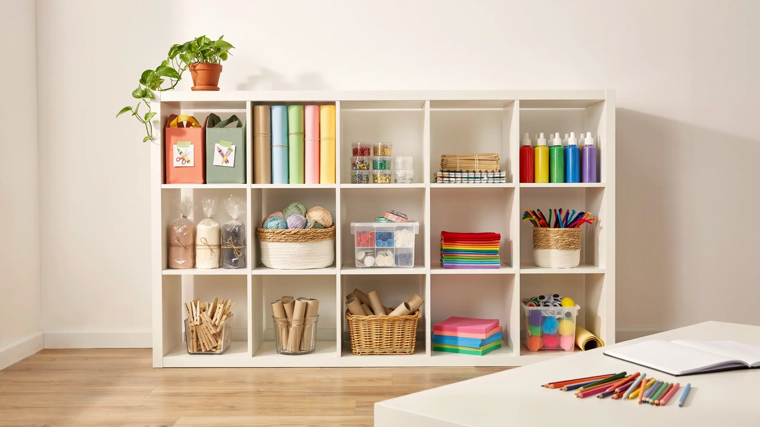 White IKEA Kallax shelving unit neatly organizing colorful craft supplies including yarn balls, paper rolls, acrylic paint bottles, and clear bead containers, with a potted green plant on top and colored pencils on a white desk in the foreground. 