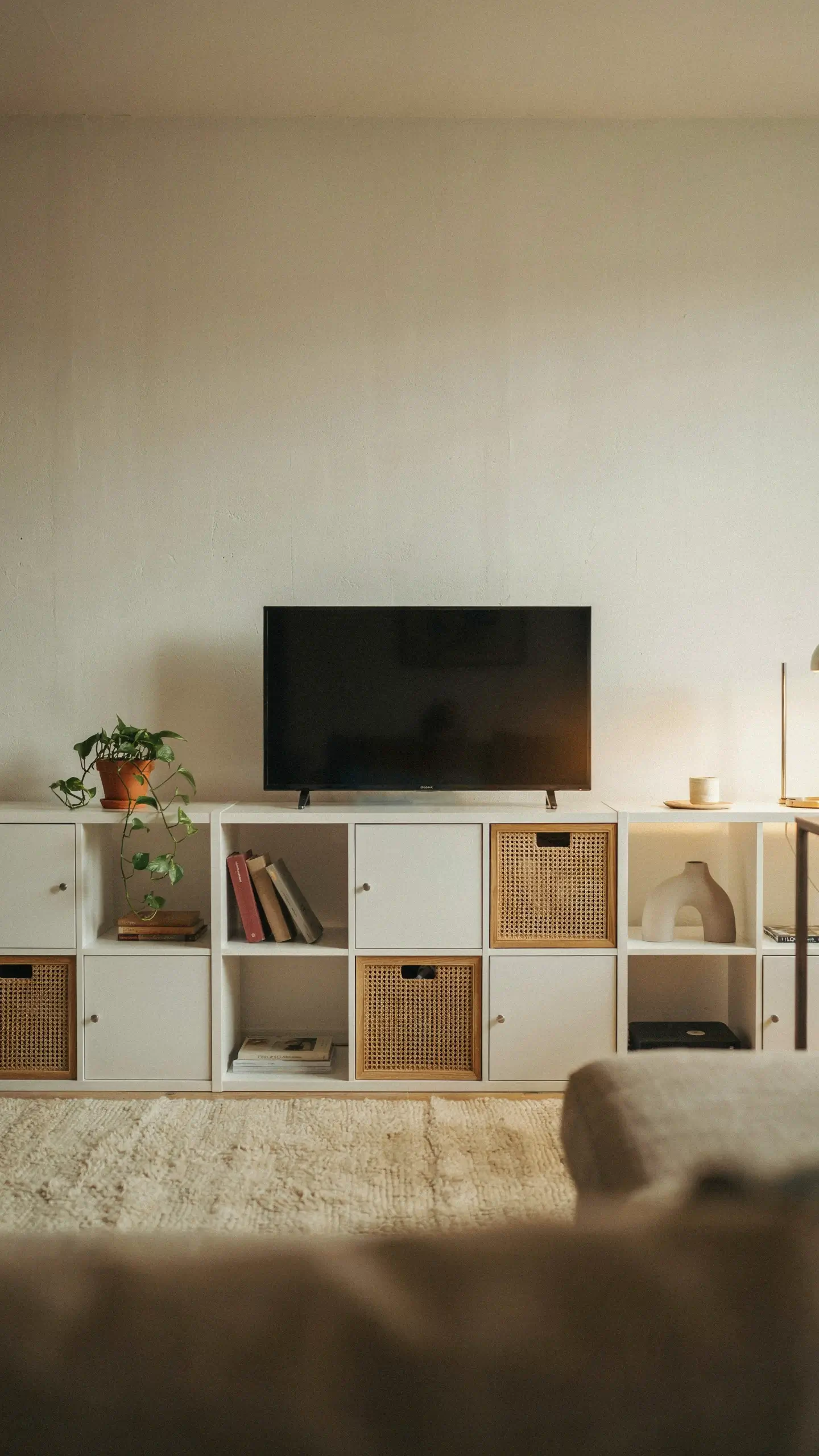 White IKEA KALLAX shelving unit styled as a TV stand with woven rattan drawers, books, and a trailing plant against a minimalist cream wall. 