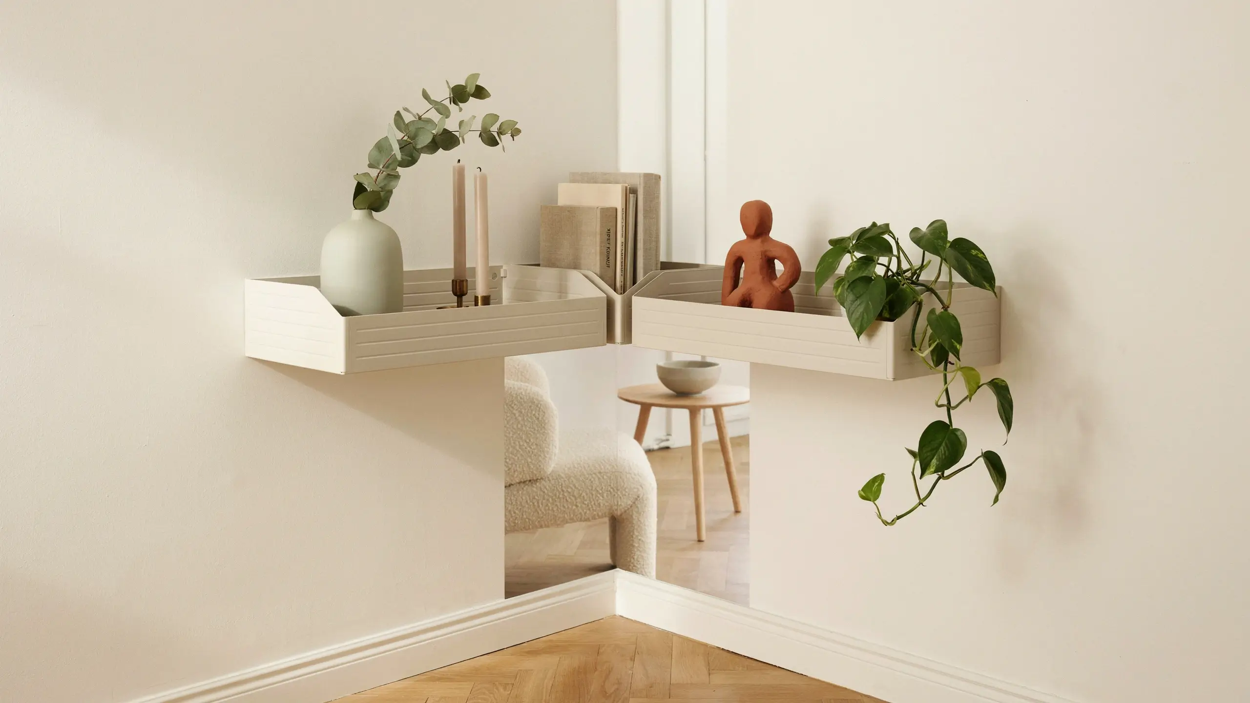 Two white IKEA KNUFF magazine file holders mounted into a wall corner to create a minimalist DIY shelf, displaying a sage green vase, eucalyptus, candles, books, a terracotta figure, and a trailing pothos plant, with a reflective mirror adding depth behind them.