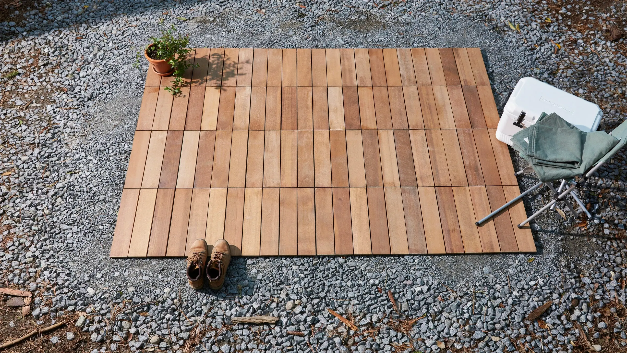 Interlocking wood deck tiles laid out on a gravel campsite to create a stable floor, surrounded by outdoor gear including boots, a potted plant, and a camping cooler and chair. 