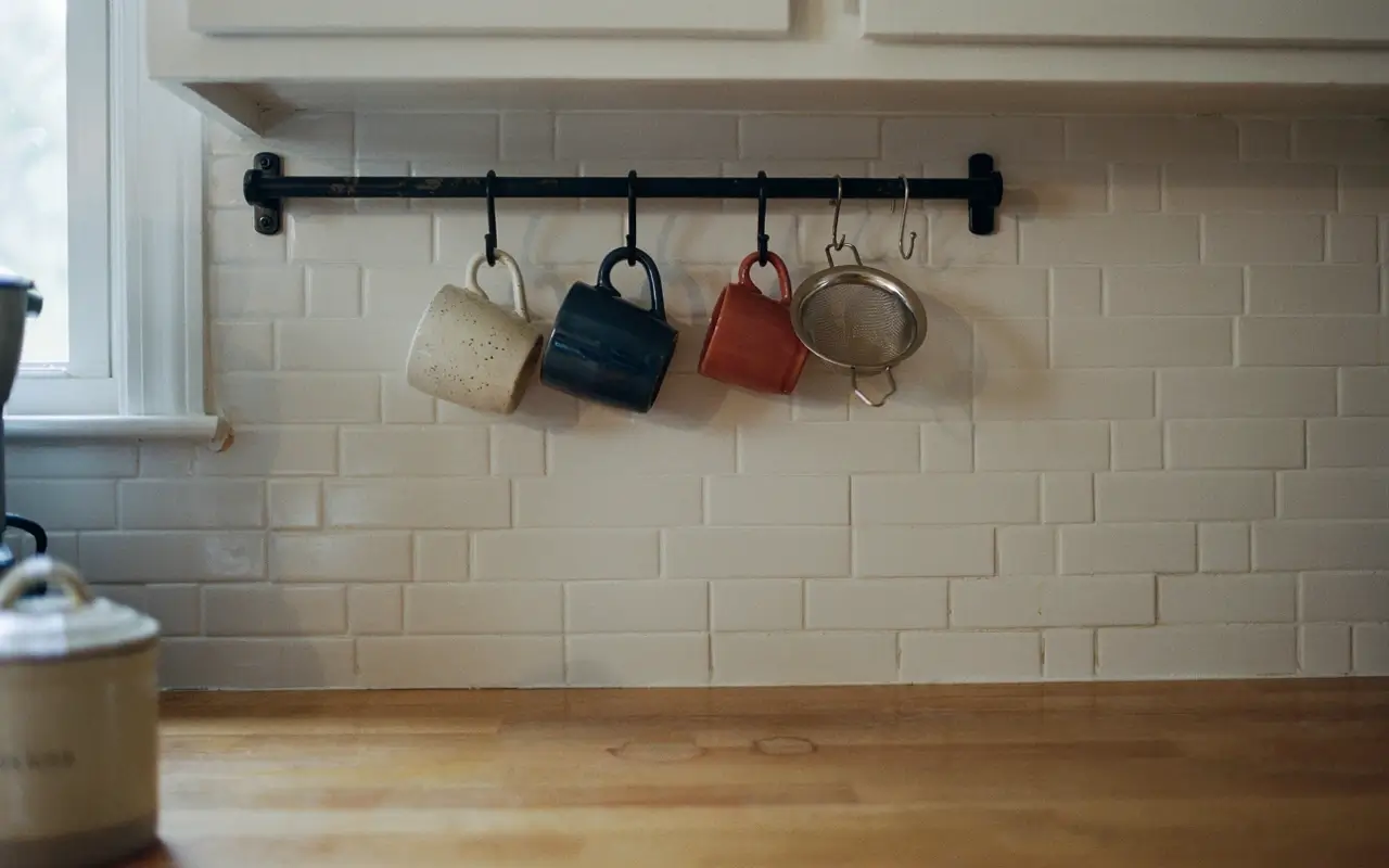 Iron utility rail with S-hooks holding mugs and a strainer under a kitchen shelf on white subway tile.