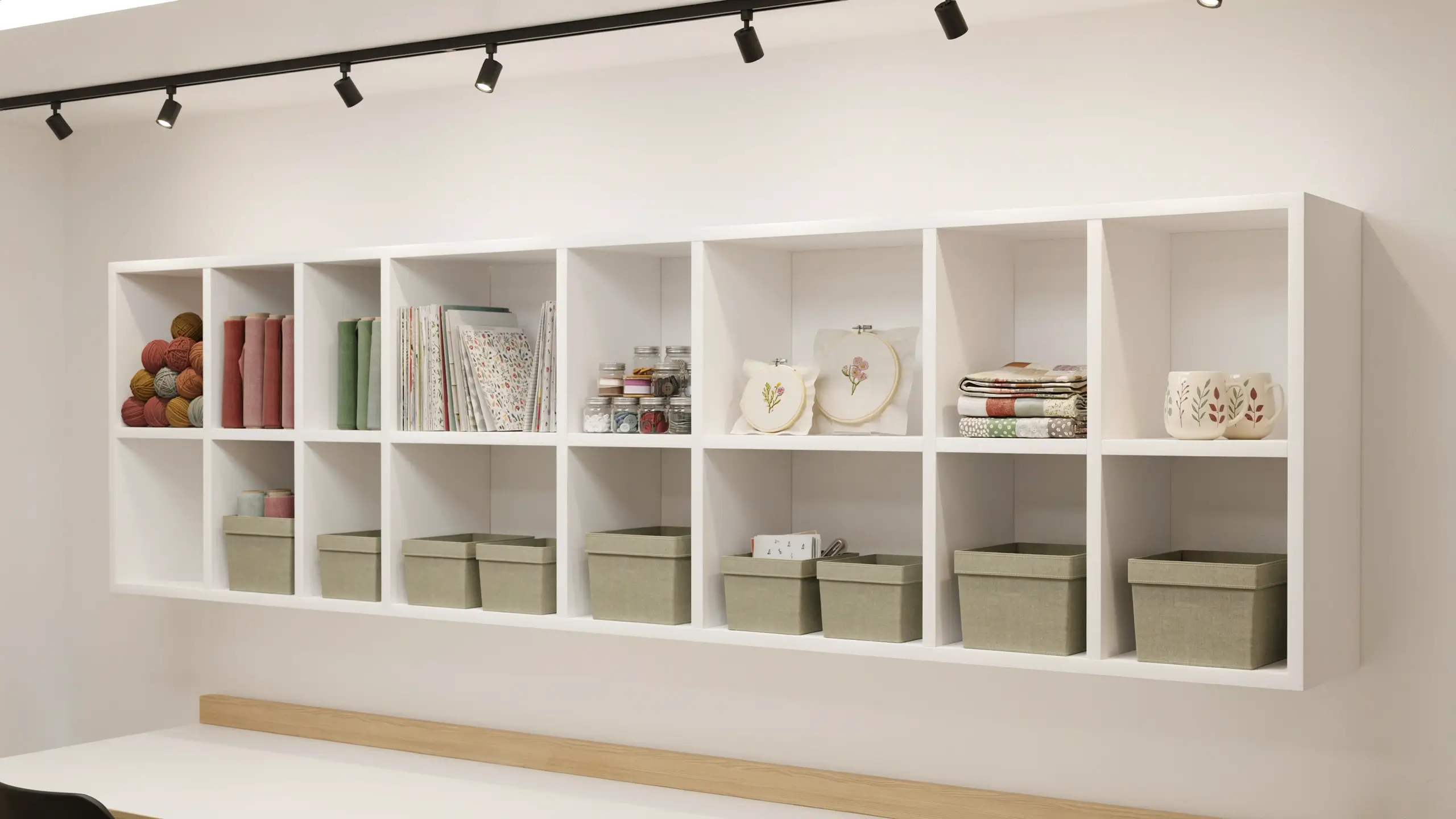 White IKEA KALLAX shelving unit used as a wall-mounted craft room organizer, displaying colorful yarn balls, binder notebooks, sewing thread jars, floral embroidery hoop art, folded fabric swatches, and matching sage green storage bins beneath black track lighting. 
