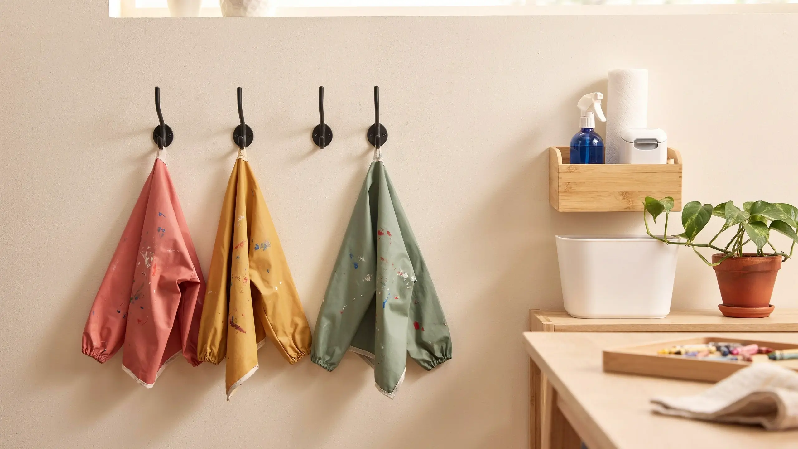"Three paint-splattered kids art smocks in pink, yellow, and sage green hanging on wall-mounted black hooks beside a bamboo cleaning shelf with a spray bottle and paper towels." 