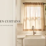 Beige linen kitchen curtains above a farmhouse sink with open shelves and trailing plant.