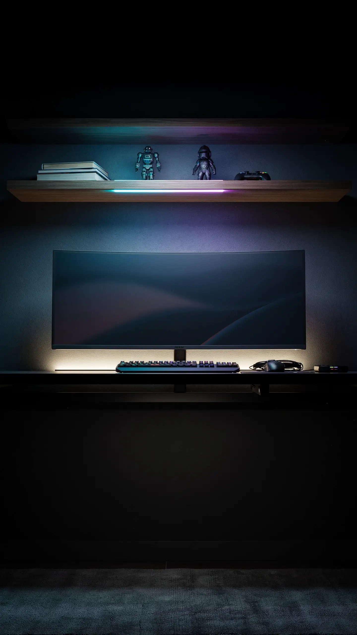 Dark minimalist gaming desk setup featuring a curved monitor, keyboard, and mouse, enhanced by a vibrant cyan and purple LED bias lighting glow beneath a floating shelf with decorative robot figures. 