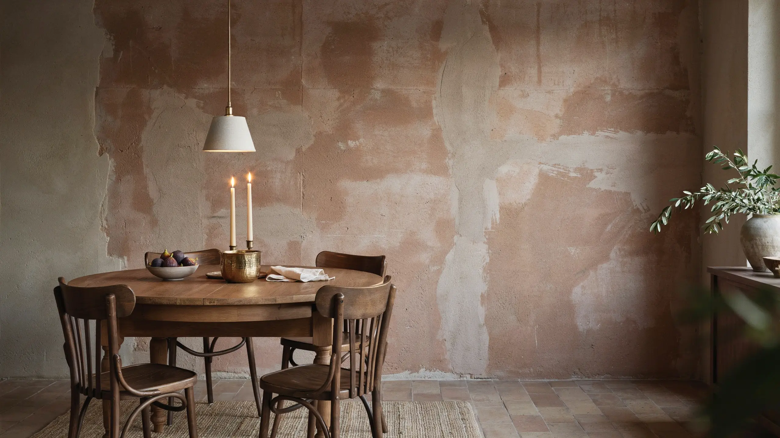 Warm rustic dining room with a textured limewash wall, dark wood oval table, spindle chairs, and a brass candle holder with figs. 