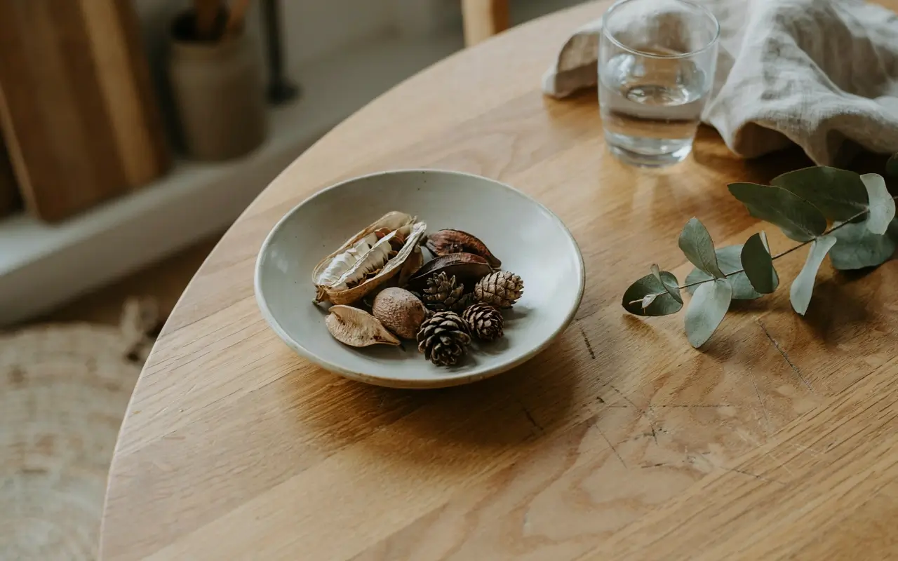 Low ceramic bowl filled with dried pods and pinecones on a round wooden table.