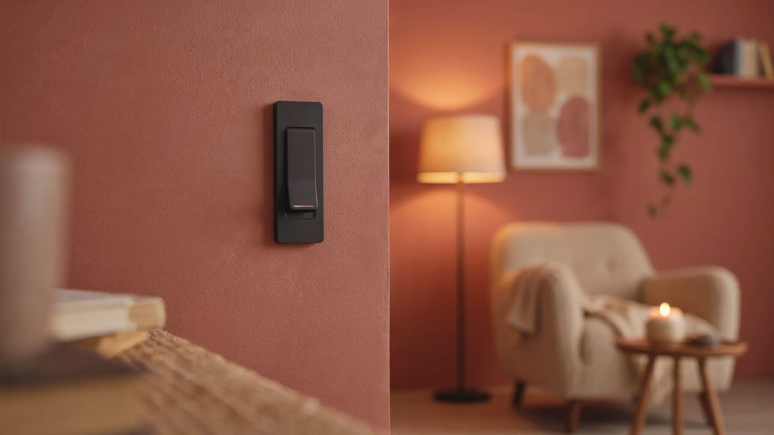 Modern matte black rocker dimmer switch mounted on a textured terracotta-painted wall, set against a blurred background of a warm-lit cozy living room with a beige armchair and a floor lamp.