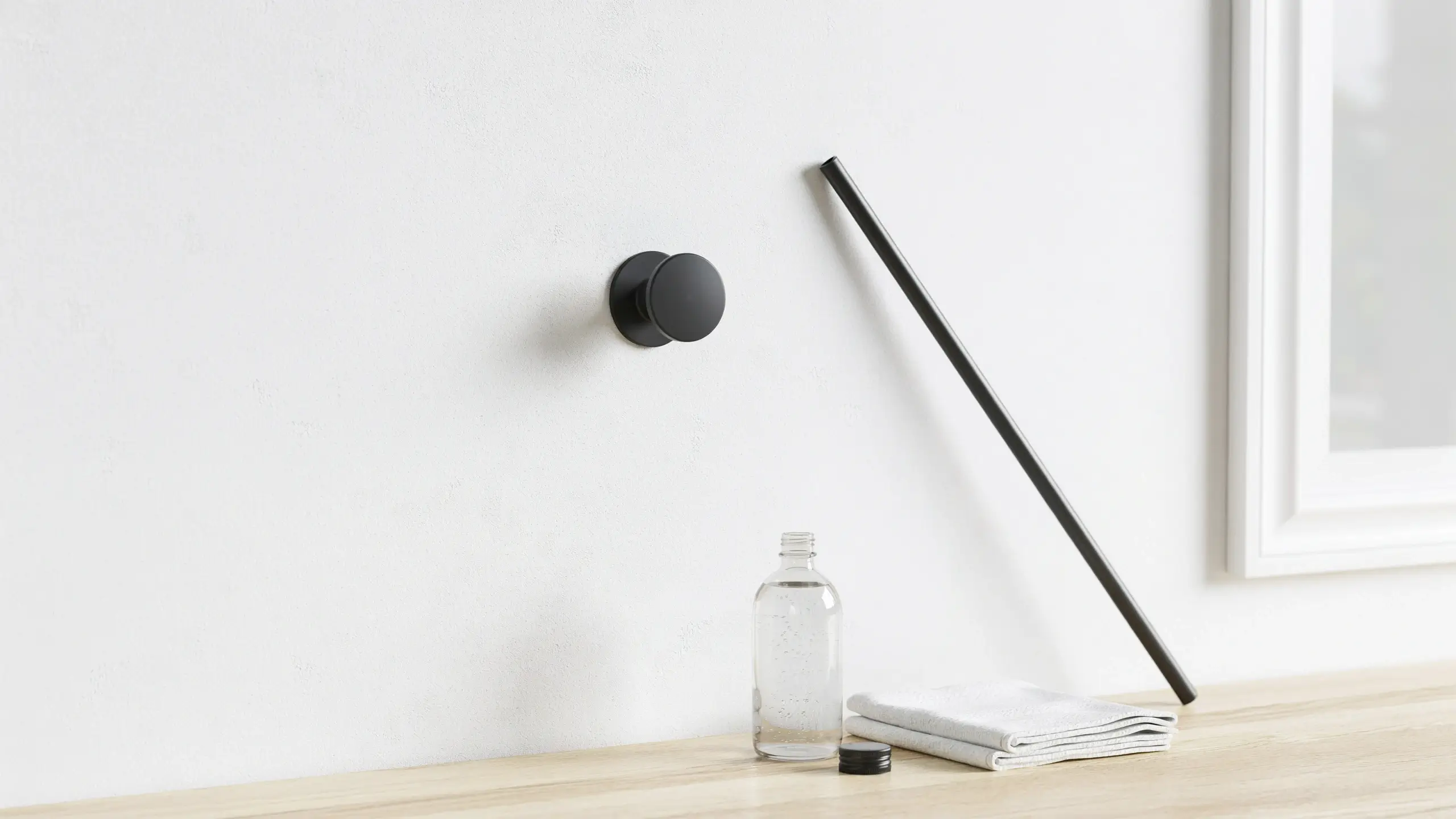 Minimalist bathroom vanity setup showing a matte black circular wall hook and angled towel bar mounted on a white textured wall, alongside a clear glass bottle and folded white towel on a light wood countertop.